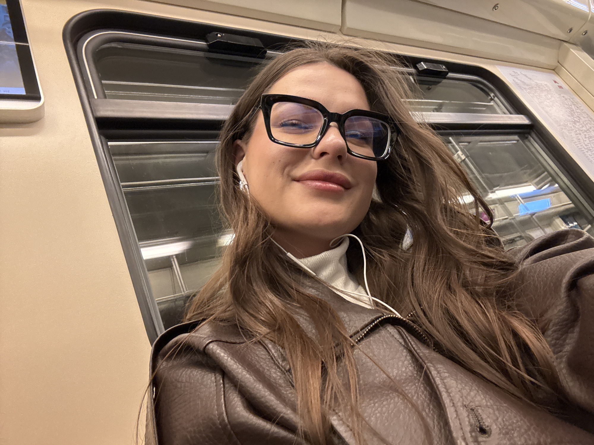A young woman with long brown hair, wearing black-framed glasses, white earbuds, a white turtleneck sweater, and a brown leather jacket, is taking a selfie inside a Moscow metro train. The interior of the train features a light beige wall, a window with multiple horizontal bars, and an information screen.