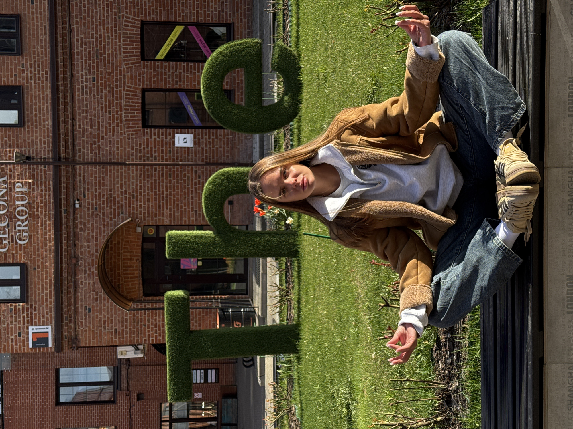 The image captures a young woman posing on a bench in front of the "OFFICE" sculpture, which is located in Moscow, Russia. The sculpture is made of trimmed green grass. She wears a tan sherpa jacket over a gray hoodie, paired with denim jeans and tan sneakers with white socks. The bench is a dark color with vertical slats. The "OFFICE" sculpture is situated in front of a brick building with several windows. The building's sign reads "GECOMA GROUP." The area is landscaped with grass and small rose bushes. The text "SHANGHAI NEW YORK LONDON" is seen on the edge of the bench.