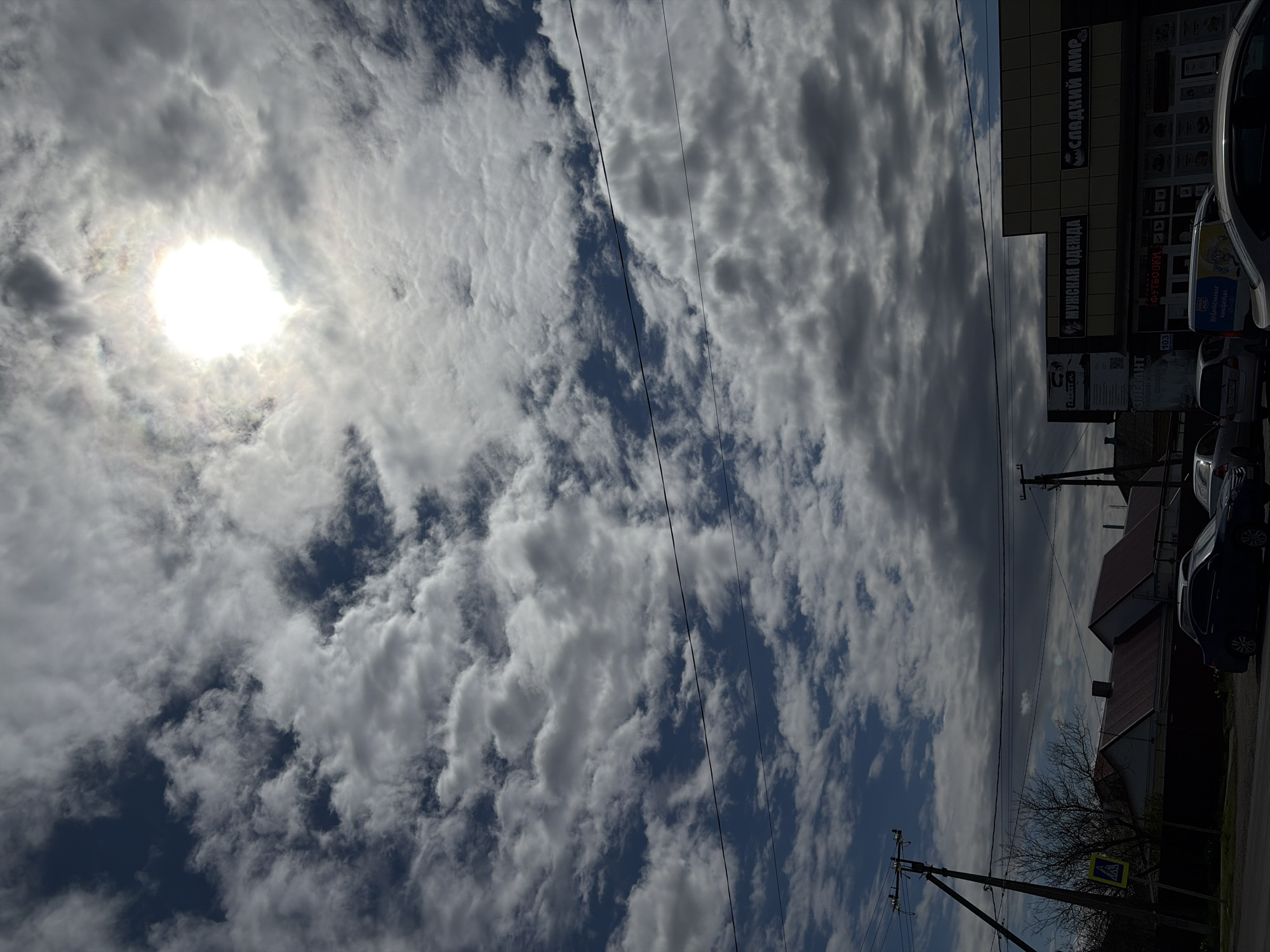 The image depicts a daytime view of a street in Unknown, Unknown. The sky is dominated by a mix of white and gray clouds, with the sun visible through a break in the clouds on the left side of the image. Power lines stretch across the sky, partially obscuring the view. On the right side of the image, there is a building with signs written in Cyrillic, including "Сладкий Мир" (Sweet World) and "Мужская Одежда" (Men's Clothing). Several parked cars are visible in front of the building. Further down the street, there are houses with red roofs and a pedestrian crossing sign. There is also a leafless tree near the street. The scene appears to be a typical urban environment.