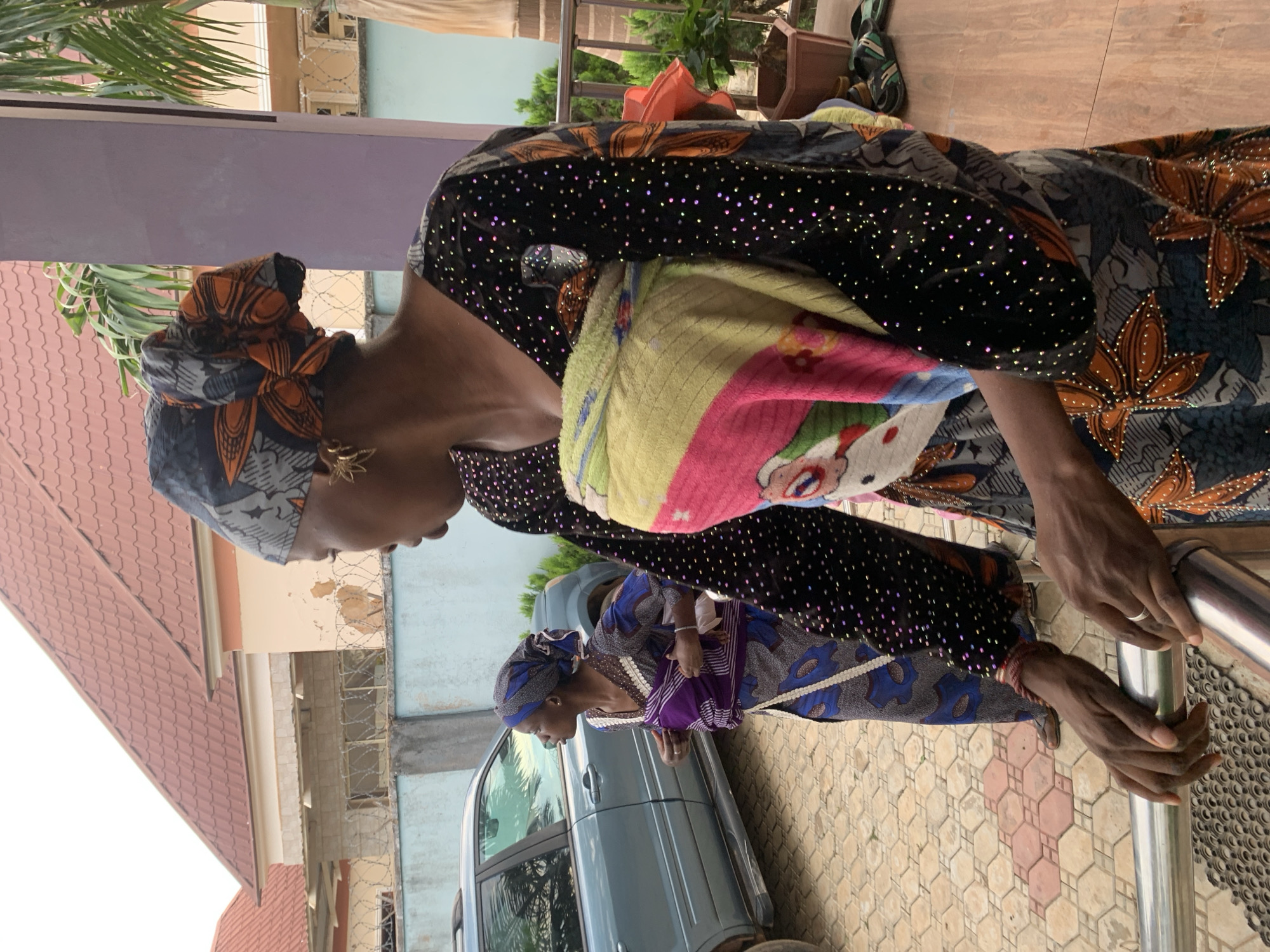 The image depicts an outdoor scene in Jos, Nigeria, featuring two women. In the foreground, a woman of dark complexion is partially visible from the chest down and the side of her face, holding onto a silver metal railing. She wears a headwrap with grey, black, and orange floral patterns and gold-colored, star-shaped earrings. Her attire consists of a dark-colored top and lower garment, both embellished with numerous small, iridescent glittery dots. A baby, wrapped in a multi-colored blanket (yellow, pink, green, white with cartoon motifs), is secured to her front. Her gaze is directed towards the left.
Behind her, slightly to the left, another woman of dark complexion stands on a paved area. She wears a blue and white patterned headwrap and a long, patterned dress in shades of blue, white, and grey, featuring brown trim around the neckline. A purple and white striped fabric is wrapped around her waist. She wears brown sandals and is also looking towards the left, with her right hand adjusting the fabric at her waist.
The ground is paved with light-colored hexagonal and square tiles, interspersed with some reddish-brown hexagonal tiles. In the background, a light blue SUV is parked. A light blue wall with barbed wire runs along the perimeter, beyond which residential buildings with reddish-brown tiled roofs and cream-colored walls, some showing signs of peeling paint, are visible. Green vegetation, including a palm tree and bushes, is scattered throughout the background. A purple pillar stands to the right of the foreground woman, and a brown plant pot containing a green plant, along with a pair of black and green sandals, are on the ground nearby. The scene is illuminated by natural daylight.
