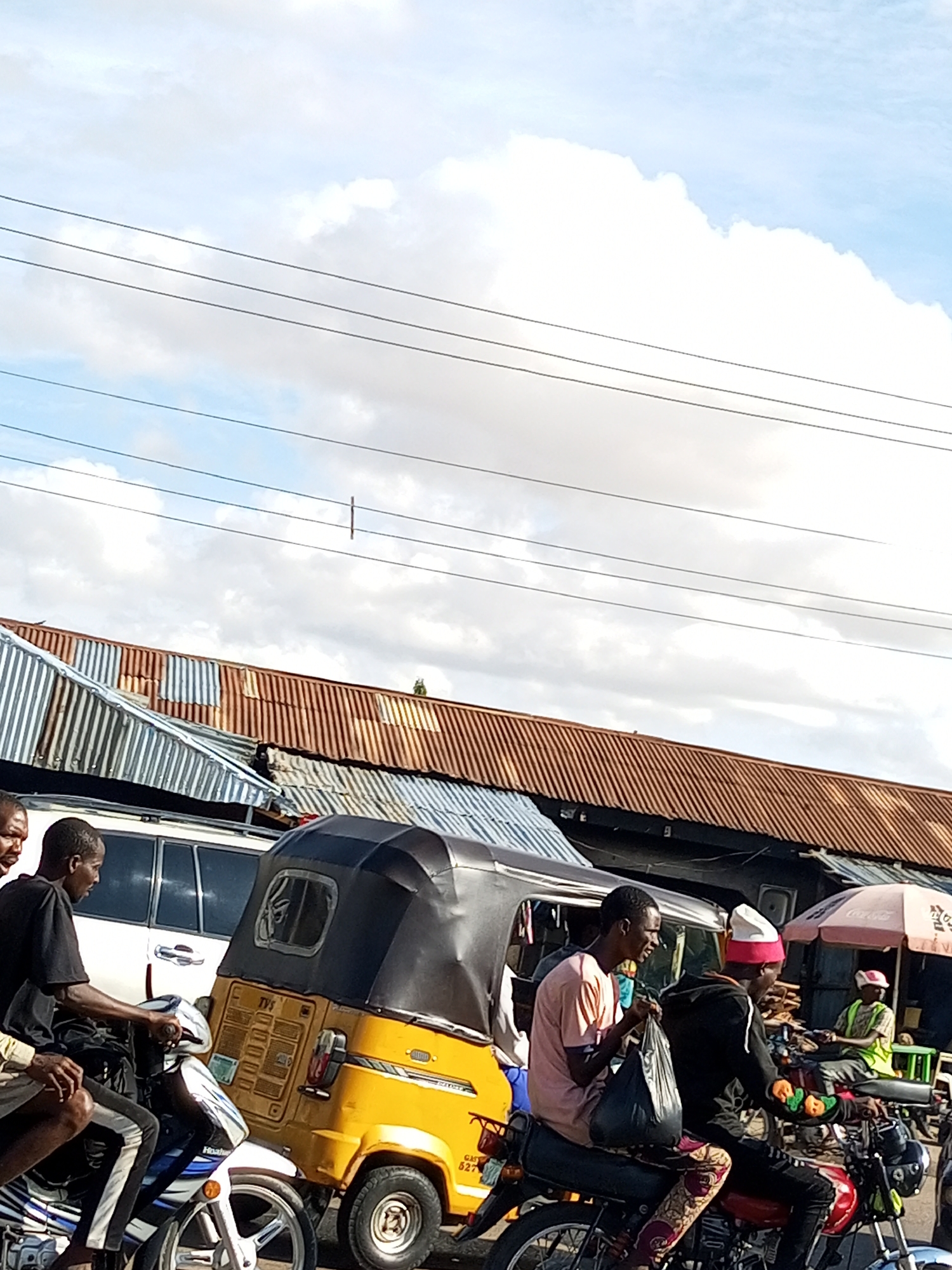 In Tumfure, Nigeria, a street scene unfolds beneath a sky filled with scattered clouds. Several individuals are present. A man in a black t-shirt rides a white and blue motorcycle. Beside him, another man in a pink t-shirt sits behind him on the same motorcycle, holding a black plastic bag. A yellow auto-rickshaw is positioned to the right of the motorcycle. Further to the right, a man wearing a white and red hat rides a red motorcycle, with a pink umbrella and a stall visible in the background. A fourth man in a green vest is seated behind the man in the red hat. In the background, corrugated metal roofs of buildings are visible. Overhead, power lines traverse the sky.