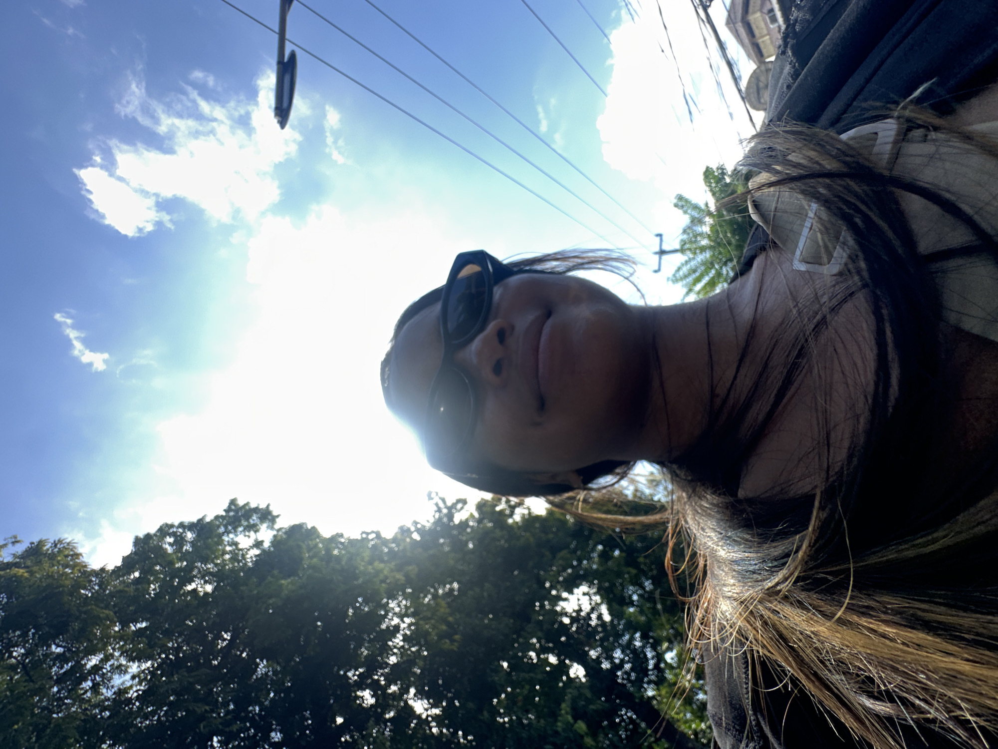 A low-angle, full-body selfie shows a young woman with dark hair and sunglasses smiling, looking up at the camera. She is outdoors, with trees and a bright blue sky with white clouds in the background. The sun is shining brightly, creating a lens flare in the image. She is wearing a dark top. The scene appears to be in a city, with power lines and a street light visible. Based on the architecture visible in the background, this image was likely taken in Denpasar, Indonesia.