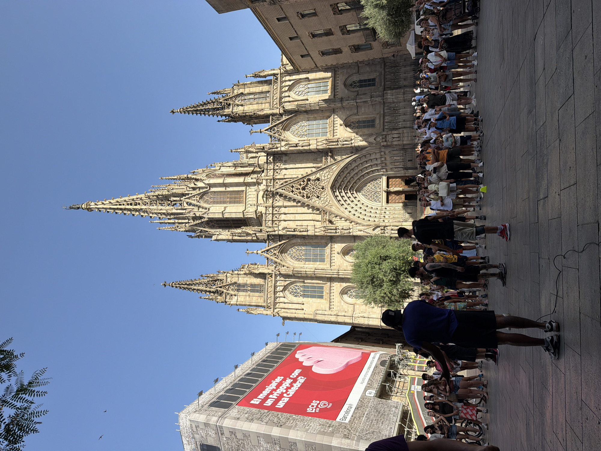 This media file captures a street scene in Barcelona, Spain, with a prominent Gothic cathedral as the backdrop. A crowd of people is gathered on a paved area in front of the cathedral, many of them standing in a line. The cathedral features intricate stone carvings, spires, and arched windows, indicative of its architectural style. The sky is a clear blue, suggesting a sunny day.
A large red billboard is visible on a building to the left of the cathedral, featuring an image of a pink ice cream bar and text in Catalan. The text reads "El menjaries un Frigopie com una Catedral?" which translates to "Would you eat a Frigopie like a Cathedral?". Below this, the words "LOCOS POR FRIGO" and "Barcelona" are visible, along with a logo. This suggests an advertising campaign for an ice cream brand.
The people in the crowd are casually dressed, wearing t-shirts, shorts, and dresses, indicating warm weather. Some are looking towards the cathedral, while others appear to be engaged in conversation or waiting. One individual in the foreground, wearing a dark blue shirt and black shorts, is facing away from the camera. The overall atmosphere appears to be one of tourism and cultural exploration, with the cathedral being a major attraction. The time of day is likely midday or afternoon, given the bright sunlight and clear sky.