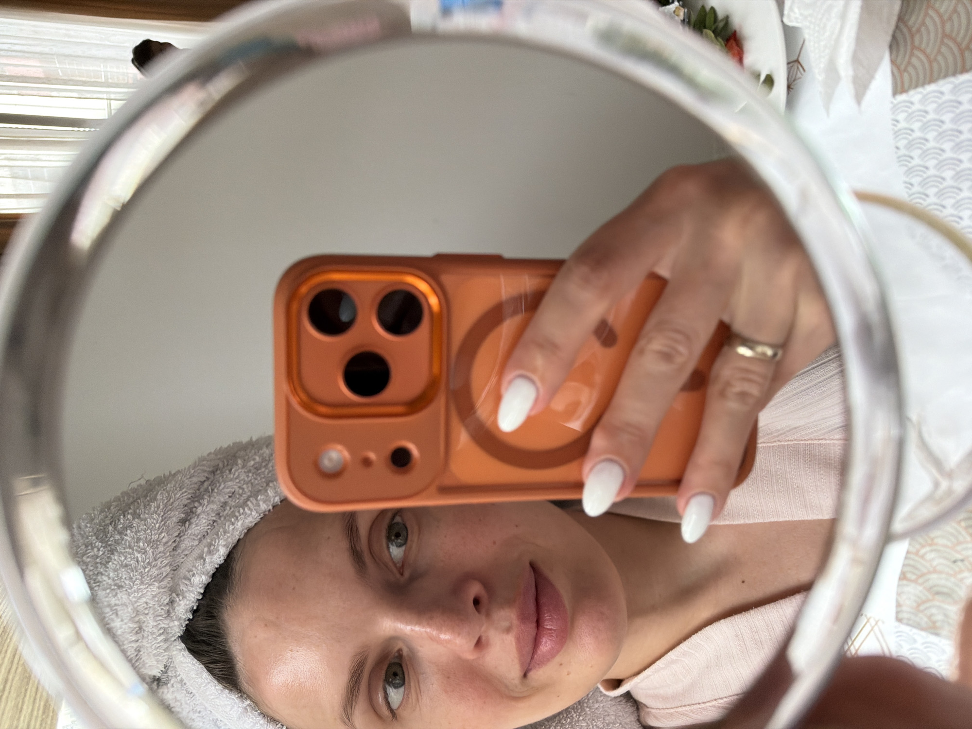 A woman is taking a selfie in a round mirror. She is holding an orange cell phone. She has fair skin, light brown hair, green eyes, and pink lips. She is wearing a gray towel on her head and a light pink top. Her nails are long and white. She is wearing a gold ring on her left ring finger. The mirror is in front of a white wall. There is a window with blinds behind the mirror and a bed with patterned bedding. The location appears to be an indoor setting. It is not possible to determine the exact location within Budva, Montenegro from this image alone.