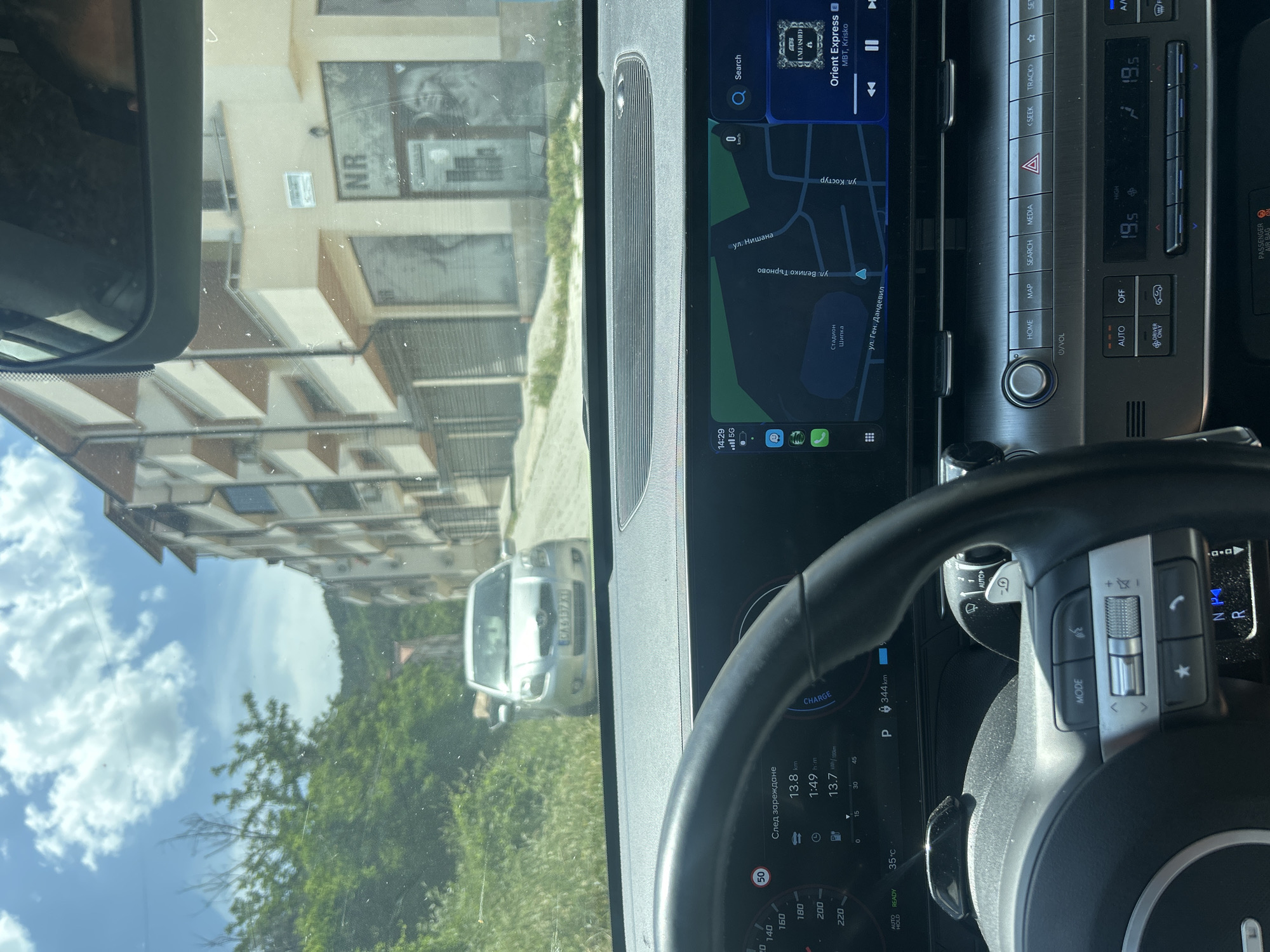 The image appears to be taken from inside a vehicle, specifically from the driver's seat, looking out through the windshield. The vehicle's dashboard and steering wheel are visible.
Through the windshield, the reflection of a building with the letters "NR" is visible, along with trees and another vehicle with license plate "CA6137XT".
The car's infotainment system displays a navigation map, showing streets like "ул. Костур" (ul. Kostur), "ул. Нишана" (ul. Nishana), "ул. Велико Търново" (ul. Veliko Tarnovo), and "ул. Ген. Дандевил" (ul. Gen. Dendevil), with a location labeled as "Стадион Шипка" (Stadion Shipka). The system also displays the song "Orient Express" by MBT, Krisko.
The car's instrument panel indicates a distance to next charge of 13.8 km, a charging time of 1:49, and 13.7 kWh/100km. The car is in Park ("P"), and the outside temperature is 35°C.