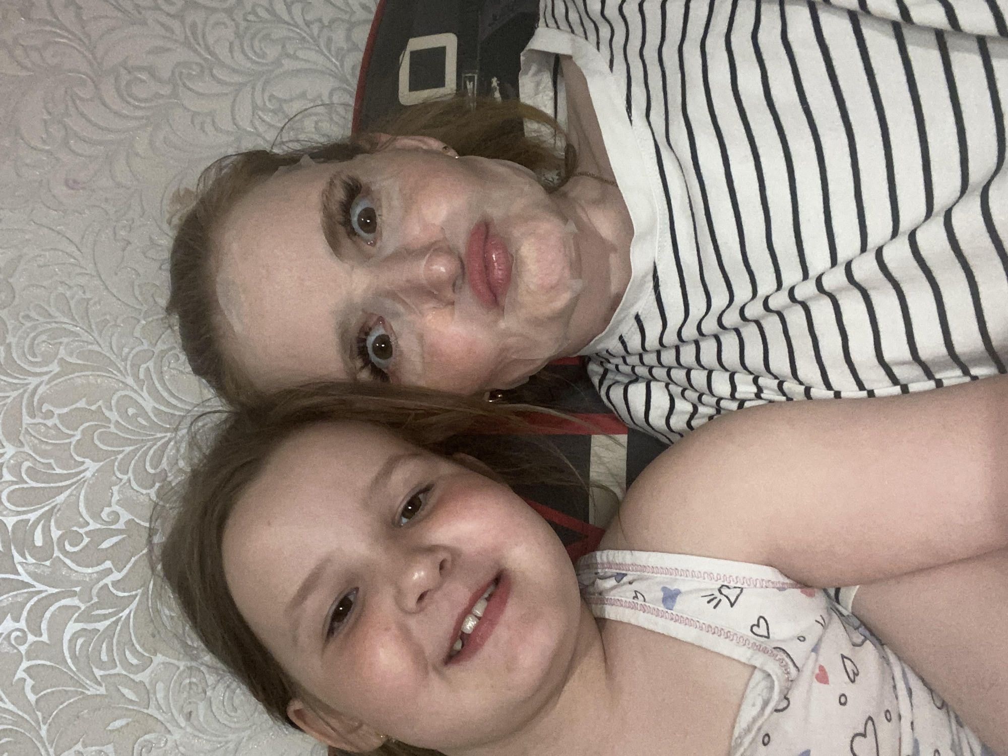 The image depicts a woman and a young girl lying on a patterned surface, likely a bed, in a domestic setting. The woman, positioned towards the top of the frame, has fair skin and appears to be wearing a facial mask. She is wearing a white and black striped shirt. The young girl, positioned below the woman, has fair skin and is smiling. She is wearing a white patterned top. The location is Chaikovsky, Russia.