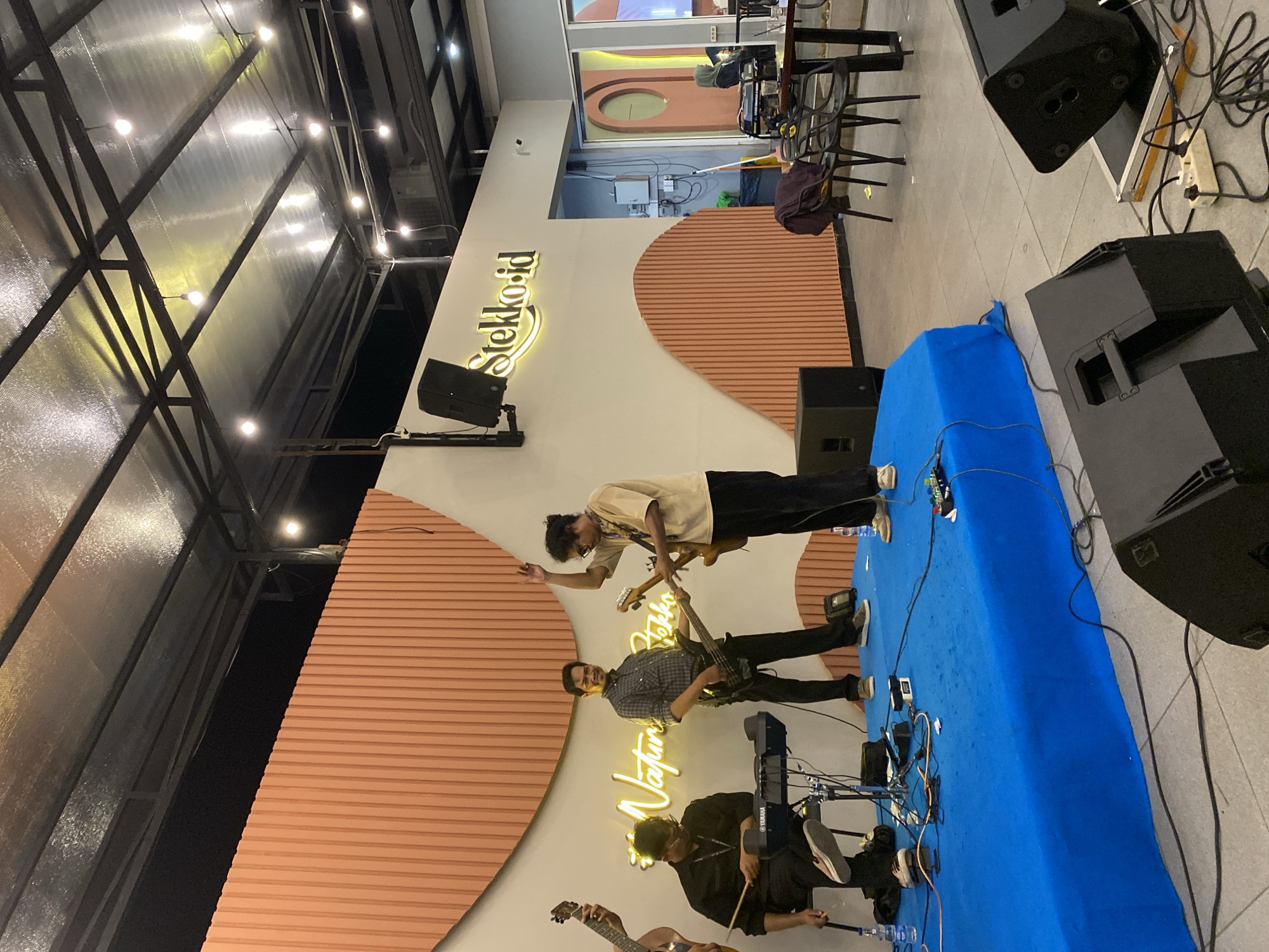 The image shows a band performing at a venue called "Stekko.id". There are three visible members: a guitarist, a bassist, and a keyboardist/percussionist. They are performing on a blue-covered stage. The venue has a modern design, with a white wall decorated with orange, half-circle patterns. Neon signs displaying the names "Stekko.id" and "Nature Park" are mounted on the wall. The location is likely in Lhokseumawe, Indonesia based on the name "Stekko.id." The ceiling has exposed metal framework and lighting fixtures. Sound equipment, including speakers, is present around the stage. Tables and chairs are visible in the background.