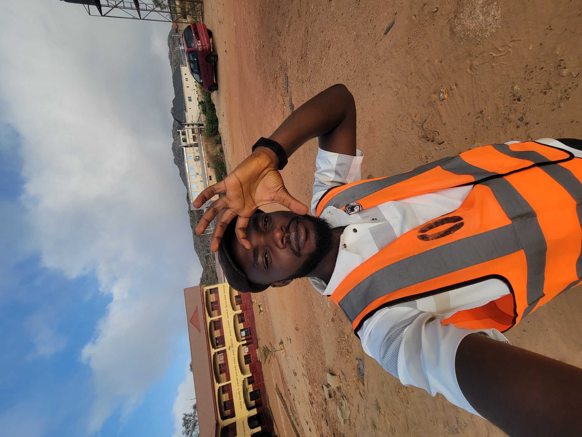 A selfie depicts a man in a bright orange and gray reflective safety vest, a white shirt with light pinstripes, and a dark-colored cap. He has dark skin, a beard, and a wristwatch. He is holding his hand up near his face.
The background includes a building with a brown roof and yellow walls, a mountainous area, a red car, a taller multi-story building, a brown dirt field, and a cloudy sky. The location is Gura Top, Nigeria.