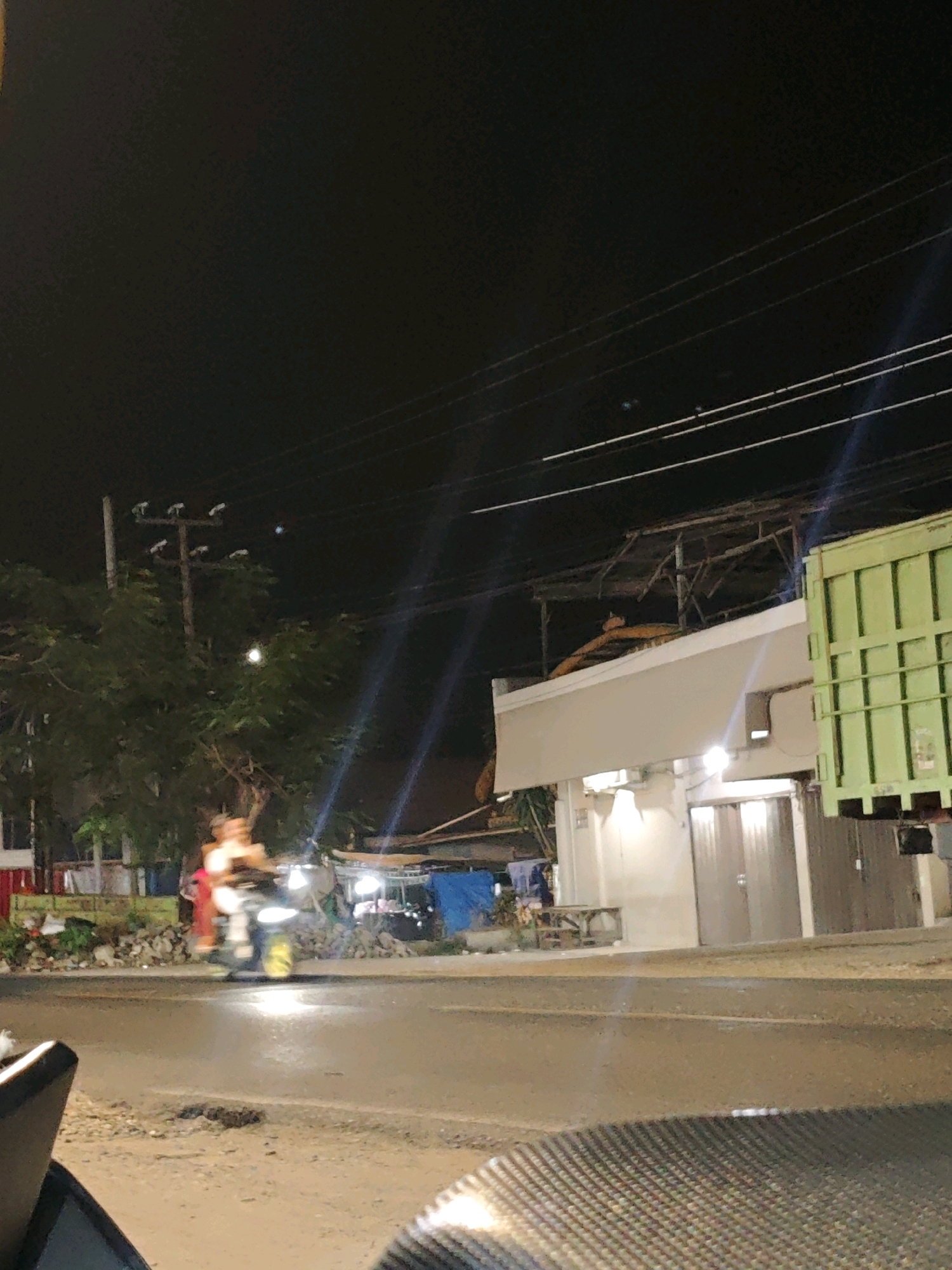 This is a nighttime, ground-level, outdoor, medium-shot photograph taken from a low angle, capturing a street scene in Gumuk Mas, Indonesia. In the foreground, the textured surface of a motorcycle's seat is visible, angled to the left. The street itself appears damp, reflecting the surrounding lights.
The midground features a busy street with traffic. A person is riding a motorcycle, moving from left to right across the frame. The rider, wearing light-colored clothing, is partially blurred due to motion. The motorcycle's headlight is illuminated, and its front wheel has yellow accents. Behind the motorcycle, a sidewalk area is visible with some debris and vegetation.
Further back, several structures line the street. On the right, a large, light green shipping container is parked. Adjacent to it is a building with a white facade and a large, gray door. The building is illuminated by lights. To the left of this building, a more rustic structure with a corrugated roof and visible scaffolding is present. In the distance, trees with lush green foliage are visible, with a utility pole and power lines overhead. The overall scene suggests a commercially active area at night, possibly with street vendors or small businesses. The sky is dark, indicating nighttime.