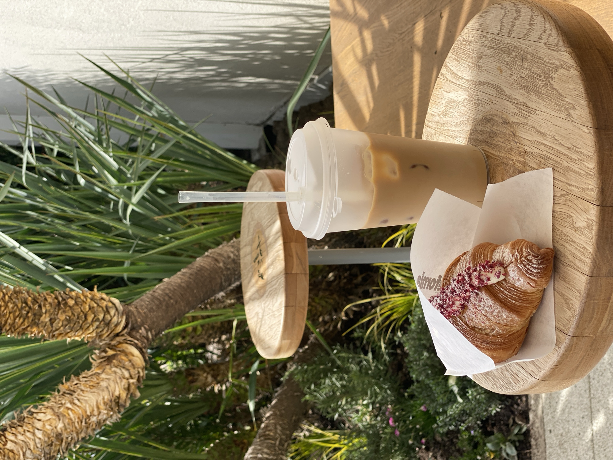 A cafe scene shows a wooden tabletop with an iced coffee in a clear plastic cup with a lid and straw. Next to the drink, a croissant dusted with a red powder sits in a white paper wrapper. The table is next to a tree and other greenery. The brand "simona" is visible on the croissant's wrapper. The scene appears to be outdoors, possibly in Sochi, Russia, based on the brand and plant life.