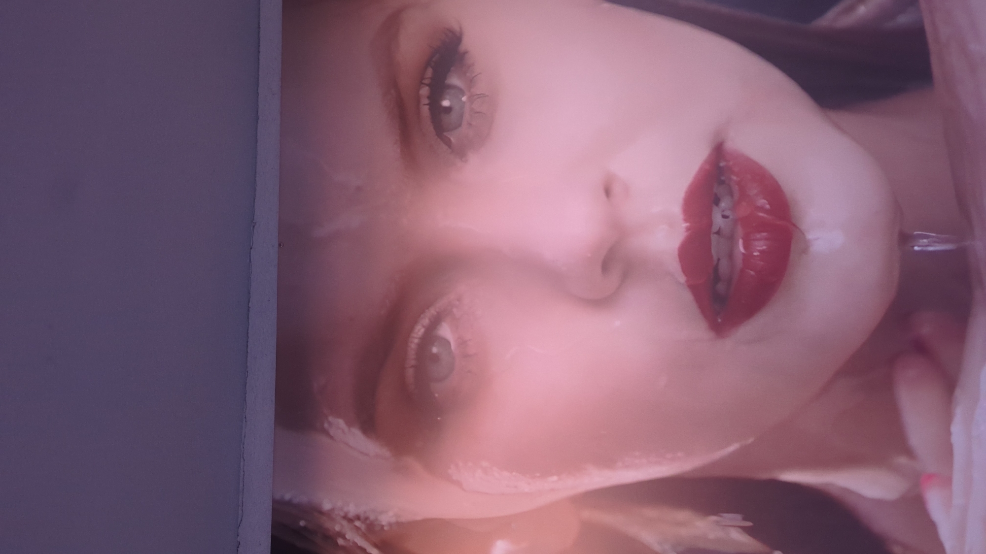 A close-up, diagonally oriented shot features a person's face, likely female, with fair skin, light-colored eyes, and bright red lipstick on partially open lips. A white or clear liquid substance covers parts of the face, including the forehead and cheek, and is visibly dripping from the right corner of the mouth. The eyes are open, and the lashes are prominent. The lighting is soft and appears to cast a warm, pinkish hue on the skin. To the immediate left of the face, a plain, light purple-grey wall or surface is visible, contrasting with the vibrant colors of the face. No text is discernible in the image.