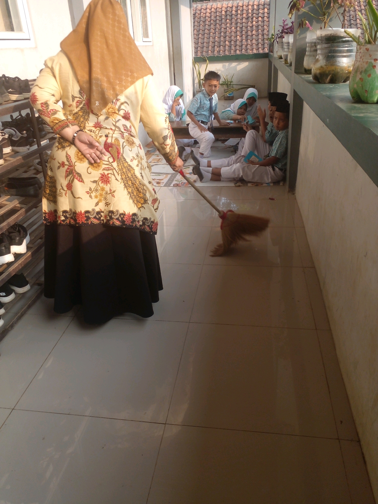 In Banyuputih, Indonesia, a woman wearing a tan hijab and a patterned yellow and black batik dress is sweeping a tiled floor with a broom. The broom's head is blurred, indicating movement. Several young boys in blue and white school uniforms are seated on the floor in the background, some appearing to read or engage in conversation. The setting appears to be an outdoor covered walkway or patio, with a wall and roof visible. Shelves filled with shoes are stacked to the left of the woman. Potted plants are situated on a ledge in the background. The lighting suggests it is daytime, with sunlight casting shadows.