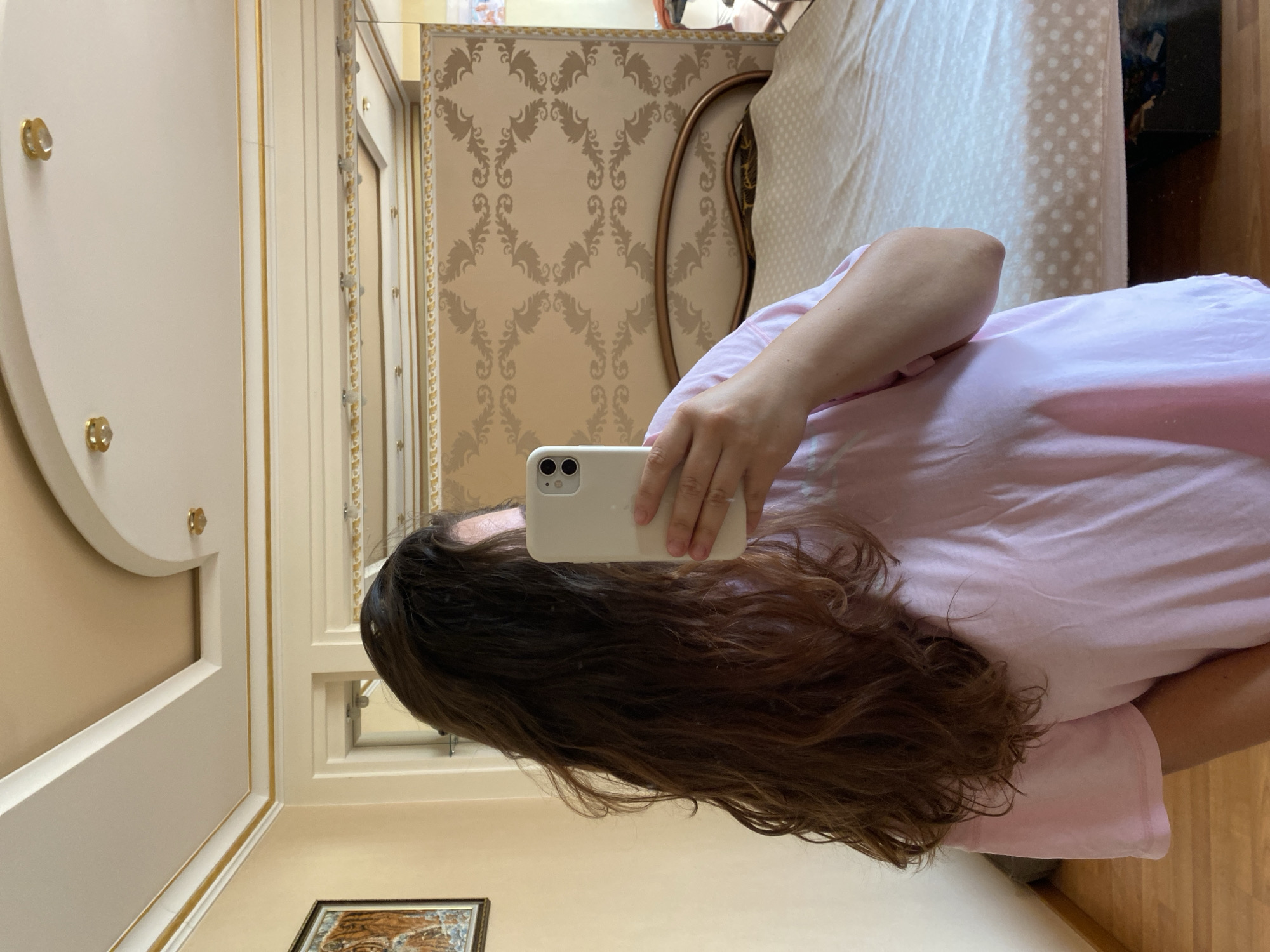 A person with long, dark brown, wavy hair, wearing a light pink t-shirt, is taking a mirror selfie in a bedroom. The person is holding a white smartphone in their right hand. In the background, a bed with a light-colored comforter is visible, along with a headboard and a patterned wall. The room appears to be decorated with ornate trim on the ceiling and walls. The image is taken in an indoor setting, a bedroom, in the city of Perm, Russia.