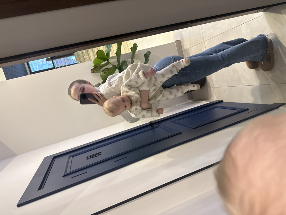 An adult female is captured in a mirror selfie, holding an infant in what appears to be a modern indoor setting within Khimki, Russia. The adult, with light-colored hair pulled back, wears a light-toned, possibly off-white, cable-knit sweater, blue flared jeans, and tan slip-on shoes. She is actively taking the photo with a dark-colored smartphone, held in her right hand, while her left hand supports the infant. A ring is visible on her left ring finger. The infant, with light hair, is dressed in a light-colored, full-body sleepsuit adorned with small heart patterns, and is looking downwards.
The environment reflects a clean, well-lit interior. To the left of the adult's reflection is a dark blue paneled door with a black handle and a black, stylized restroom symbol depicting male and female figures. The surrounding walls are white, and the floor is covered with light grey rectangular tiles. To the right of the adult, a large green houseplant with broad leaves is visible in a dark pot. Further into the background, reflected through what appears to be a large window or glass partition, is a modern building interior, potentially multi-storied, featuring structural elements and suggesting a commercial or public establishment. Based on the visual evidence, a specific address or named venue within Khimki cannot be determined.