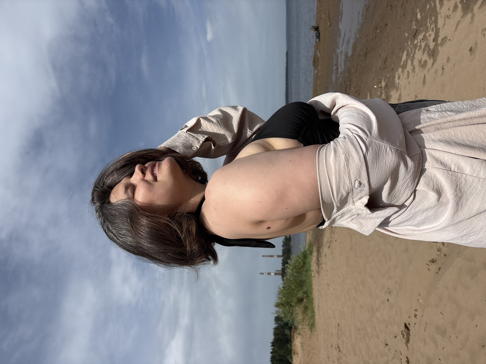The image depicts a woman on a sandy beach. She is wearing a black top and a light-colored open shirt. Her hair is dark with grey streaks. She is positioned with her back to the camera, her face visible and tilted upward. The sky is visible with scattered clouds. In the background, across the water, there is vegetation, and two industrial smokestacks can be seen, indicating a location in or near the city of Chaikovsky, Russia. The beach appears mostly empty.