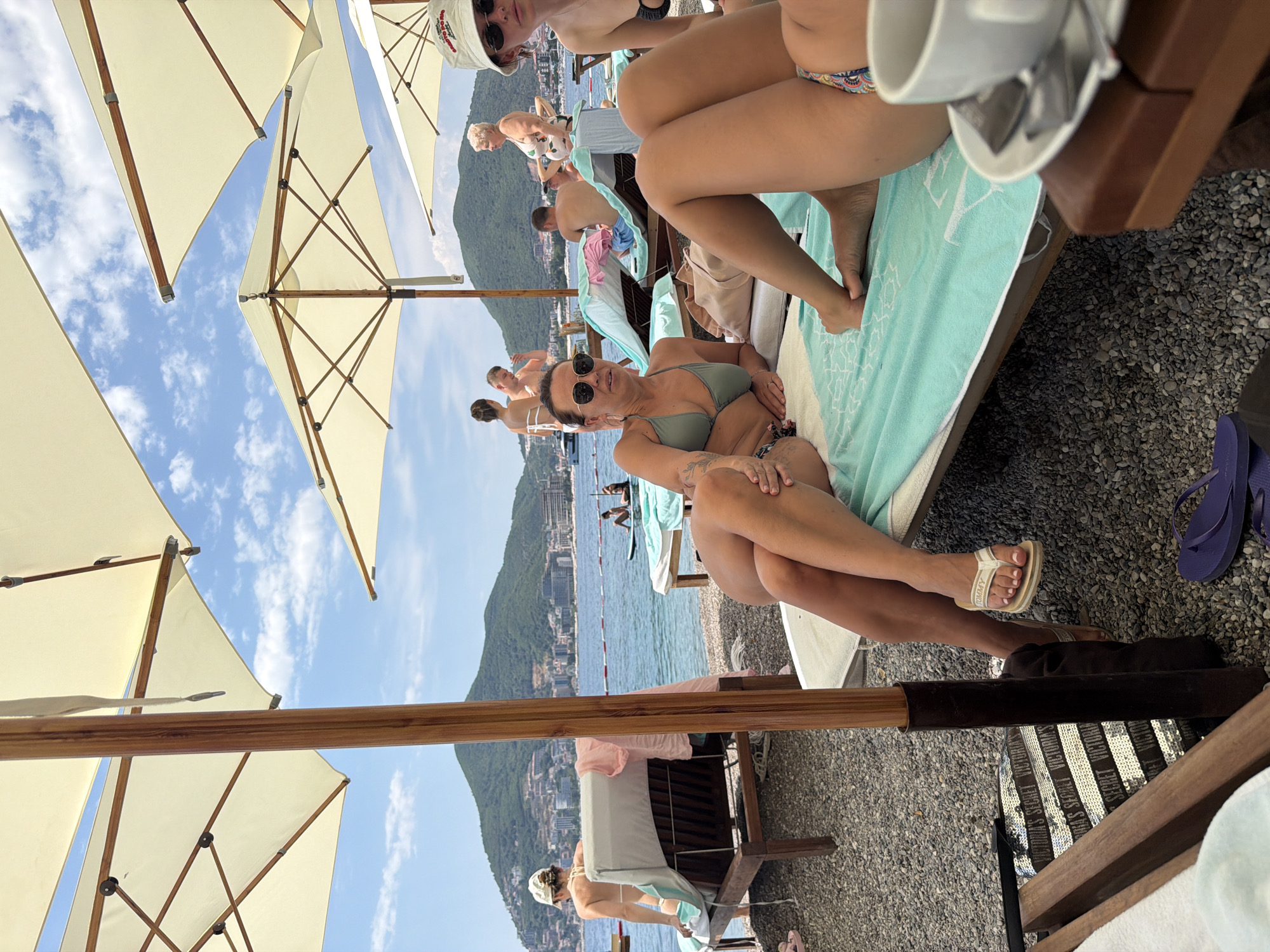 The media file shows a sun-drenched beach scene in Budva, Montenegro. The perspective is from behind a beach chair, looking towards the sea and a lush green hillside dotted with buildings. Several beach umbrellas, made of cream-colored fabric and supported by wooden poles, dominate the upper portion of the image, casting shadows on the pebble beach.
In the foreground, a woman with dark hair and sunglasses is lying on a turquoise beach towel. She is wearing a green bikini and appears relaxed. To her right, another woman's legs are visible, also on a turquoise towel. She is wearing a patterned bikini.
Further back, other people are enjoying the beach. A man in a white shirt and cap is seen in the middle ground, and behind him, several other individuals are lounging or walking. The sea is calm and blue, with a few boats visible in the distance. The hillside scenery suggests a coastal town.
The time of day appears to be sunny and warm, characteristic of a summer afternoon. The sky is a bright blue with scattered white clouds. The overall mood is one of leisure and relaxation on a beautiful vacation day. No visible text is present in the image, except for what appears to be a very small brand logo on the woman's sandal in the foreground.