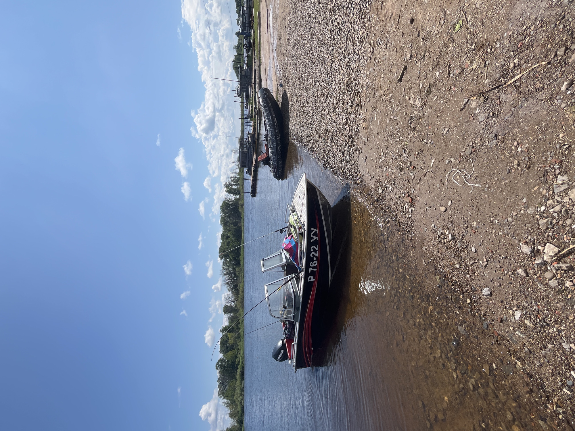This image shows a sunny day at the shore of a river in Ust-Belsk, Russia. In the foreground, a small motorboat with the registration number P 76-22 YY is partially submerged in the clear water near a rocky beach. The boat is black with red and silver accents and has an Evinrude outboard motor. Several fishing rods are visible in the boat. To the right of the motorboat, a camouflage-patterned inflatable boat is resting on the shore. In the background, several other boats are docked on the river, and a line of trees borders the opposite bank. The sky is blue with scattered white clouds.