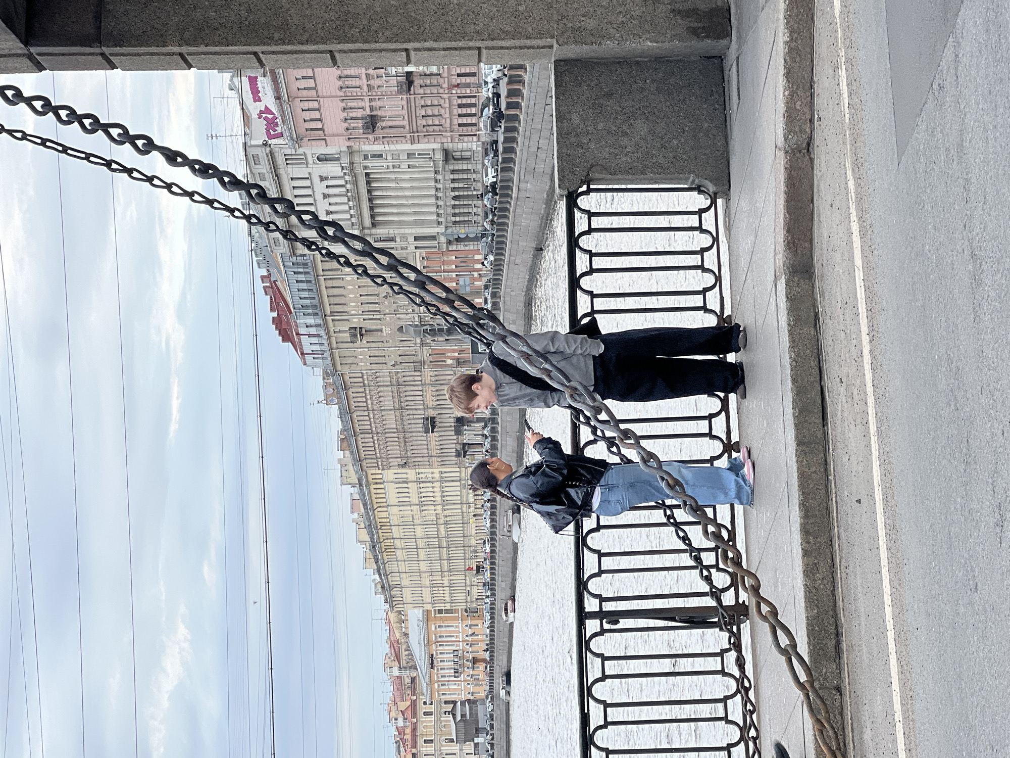 The image, when viewed correctly oriented, depicts two individuals standing by an urban canal or river in Saint Petersburg, Russia. The scene is captured from a slightly elevated position, possibly from beneath an archway or bridge, with heavy, dark metal chains prominent in the foreground, hanging diagonally across the left side of the frame. These chains are characteristic features of the city's bridges and embankments.
A man, dressed in a grey hoodie and dark trousers, stands closer to the foreground, looking downwards towards the water. Beside him, a woman wearing a black leather jacket, light blue jeans, and pink shoes is holding a smartphone, seemingly pointing it towards the water or the opposite bank, possibly taking a photo or video. Both are positioned behind a black, decorative metal railing that separates the paved walkway from the water.
Across the calm water of the canal or river, a long stretch of multi-storied, classical-style buildings lines the opposite embankment. A road with several cars is visible running parallel to the water. The sky is overcast, indicating a cloudy day. Faint power lines are visible against the sky in the distance. On one of the more distant buildings across the water, faint text "ZOOBA", "РЯДОМ" (Russian for "nearby"), and partially "БОЛЬШАЯ" (Russian for "big" or "large") can be discerned, likely as signage or graffiti. The overall setting is an urban landscape with historical architecture.