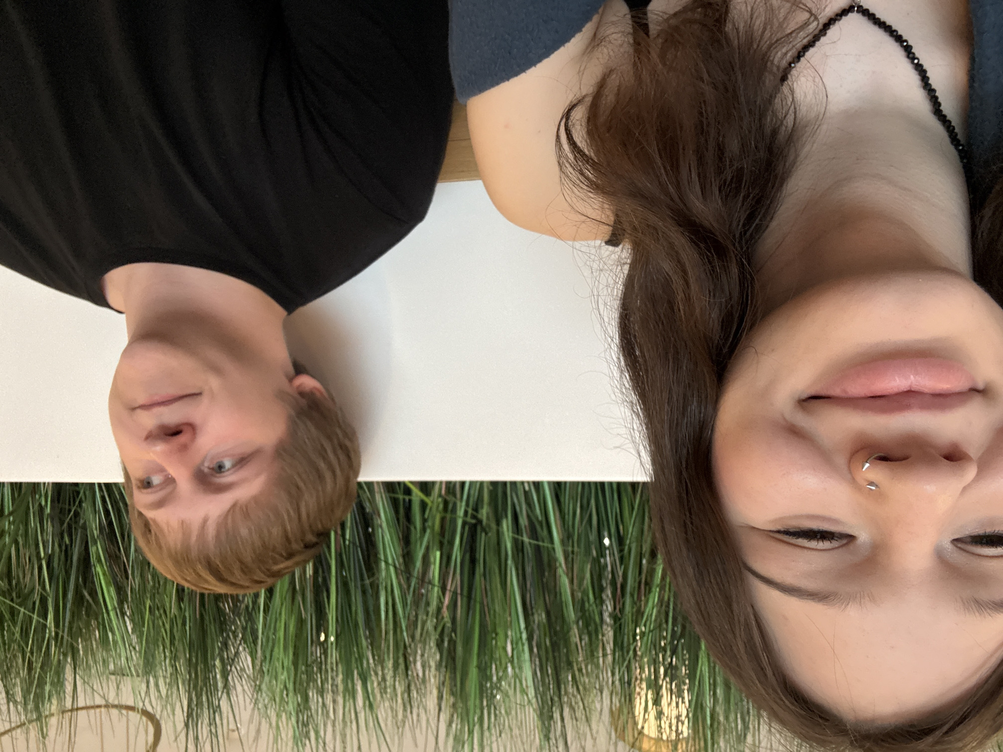 This image depicts two individuals seated in an indoor setting, contextually located in the city of Saint Petersburg, Russia. The subject on the left, a female with medium-long dark brown hair, is positioned closer to the camera. Her face shows a smiling expression with eyes mostly closed, and she wears a silver spiral nose ring featuring two small spikes. A black beaded necklace with a small pendant is visible around her neck, and she is wearing a dark garment, possibly a top, beneath a dark blue fabric draped over her left shoulder. The subject on the right, a male with short light brown hair, is seated further back, looking upwards and to his right with a neutral to slightly smiling expression. He wears a plain black crew-neck t-shirt. Both individuals are fair-skinned. They are seated in front of a white wall or barrier. Behind this barrier, a dense arrangement of artificial green grass-like foliage extends across the background. Above the foliage, glimpses of a ceiling or upper wall structure are visible, featuring what appears to be a light-colored surface and a curved, golden-toned architectural element, possibly part of a light fixture. A dark wooden surface is visible at the bottom right, possibly indicating a table or bench.