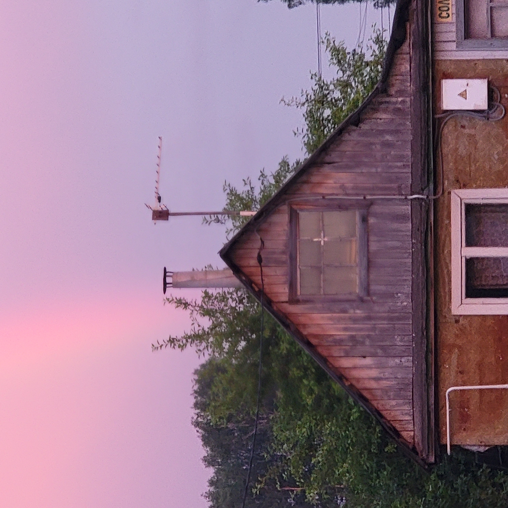 The image depicts the upper portion of a weathered, two-story building against a soft, gradient sky of pink and purple hues. The building features a prominent gable roof constructed of vertical wooden planks. A small, frosted window with a cross-shaped divider is visible in the gable. Below the gable, the first story appears to be made of rough, textured material, possibly stucco or concrete, with a window featuring a light pink frame and divided panes. A white pipe extends horizontally along the top of the first story.
On the roof, a grey, cylindrical chimney stands to the left, and an old television antenna is mounted on a pole to the right. Green foliage from trees surrounds the building, with some branches reaching up towards the roof. The sky suggests either dawn or dusk. On the right side of the building, a white electrical box with a yellow triangle symbol is affixed to the wall, and a yellow sign with Cyrillic lettering, partially visible as "СОМ", is present. The scene is likely a residential building in Iglino, Russia, given the context. No people or animals are visible in the image.