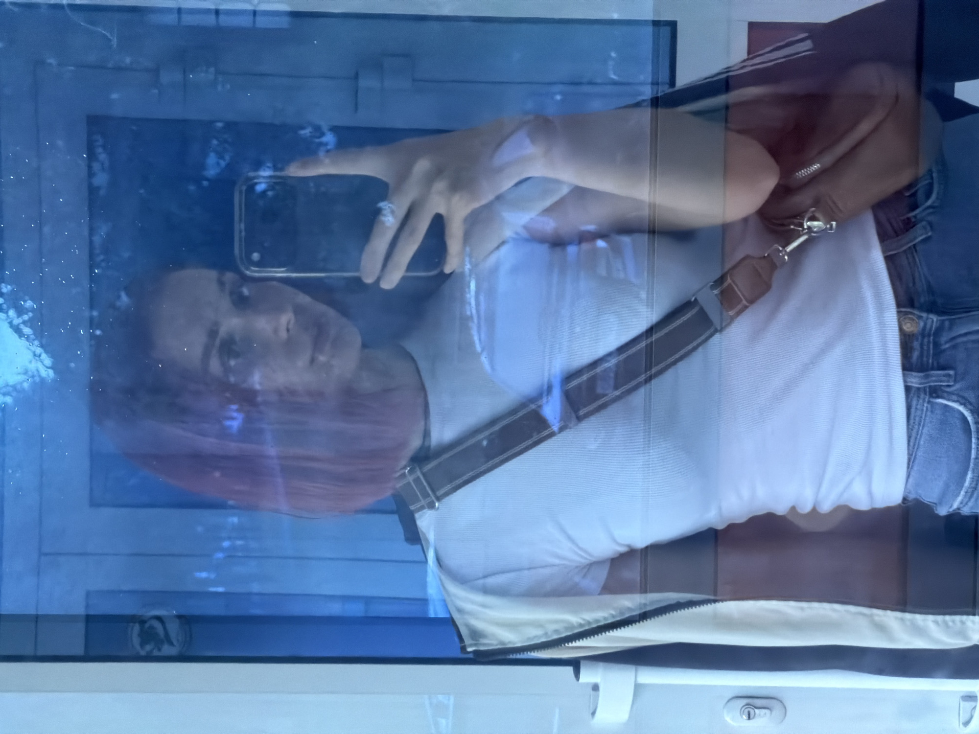 A woman with reddish-brown hair is taking a mirror selfie. She is wearing a white ribbed t-shirt, a light-colored jacket zipped down, and blue denim jeans. A brown crossbody bag with a striped strap is slung across her shoulder. She is holding a smartphone in her right hand, with a ring visible on her ring finger. The background appears to be a reflective surface, possibly glass, with a blue tint and some reflections of what could be doors or panels. A white door frame and a door lock are visible to the left of the woman. The overall lighting is dim, with some bright reflections. The scene is set indoors, likely in a hallway or room with a reflective surface. There are no clear indicators of the specific location within Chaikovsky, Russia, based solely on the visual information provided.