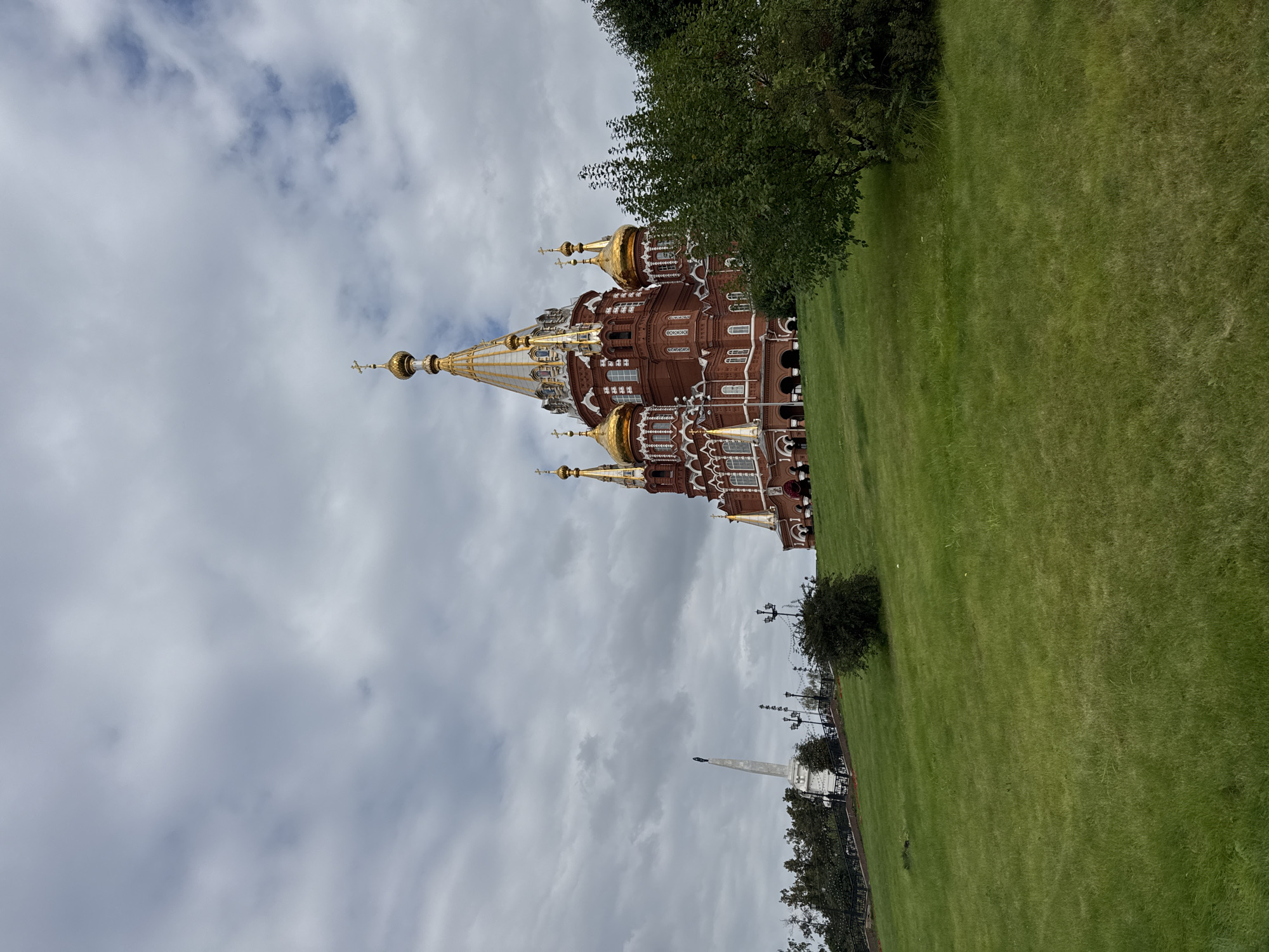 The image shows the Alexander Nevsky Cathedral in Izhevsk, Russia, on a cloudy day. The cathedral is a large, red brick building with multiple golden domes and spires. It is situated on a grassy hill, and in the foreground, there is a large expanse of green grass. To the left of the cathedral, there is a tall, white obelisk monument. Trees are visible on the left and right sides of the image, and lampposts line a pathway leading up to the cathedral. The sky is filled with dramatic, gray clouds. There are no people visible in the image.