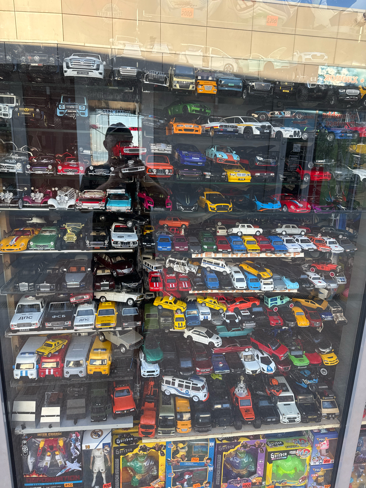 The image shows a display case filled with numerous toy cars and action figures in a shop in Anapa, Russia. The display case is made of glass and is illuminated from within, reflecting the surrounding environment and a person's silhouette, likely a customer or the shopkeeper.
The toy cars are arranged in rows on multiple shelves. There are various types of toy cars, including SUVs, sedans, sports cars, trucks, and vans, in a multitude of colors such as white, black, red, blue, yellow, green, and silver. Some of the cars are detailed replicas of real vehicles, while others appear to be more generic models. Several cars have visible branding or license plates with Russian text, such as "ДПС" (D.P.S. - State Traffic Inspectorate) and "ФСБ" (FSB - Federal Security Service).
In the lower section of the display, several action figures and boxed toys are visible. These include transformers, characters from popular franchises, and stretchy toy figures. One prominent box features a stylized character with the text "STRETCH" and "ROCK JAY". Another box displays a figure of a football player, likely Cristiano Ronaldo, wearing a Real Madrid uniform.
The background visible through the glass suggests an indoor setting with tiled walls and some signage, including what appears to be price tags or advertisements with numbers like "2000". The overall scene is that of a toy store or a stall selling collectible model cars and toys. The lighting is bright, creating reflections on the glass surface. The image is taken from a slightly elevated angle, looking into the display case.