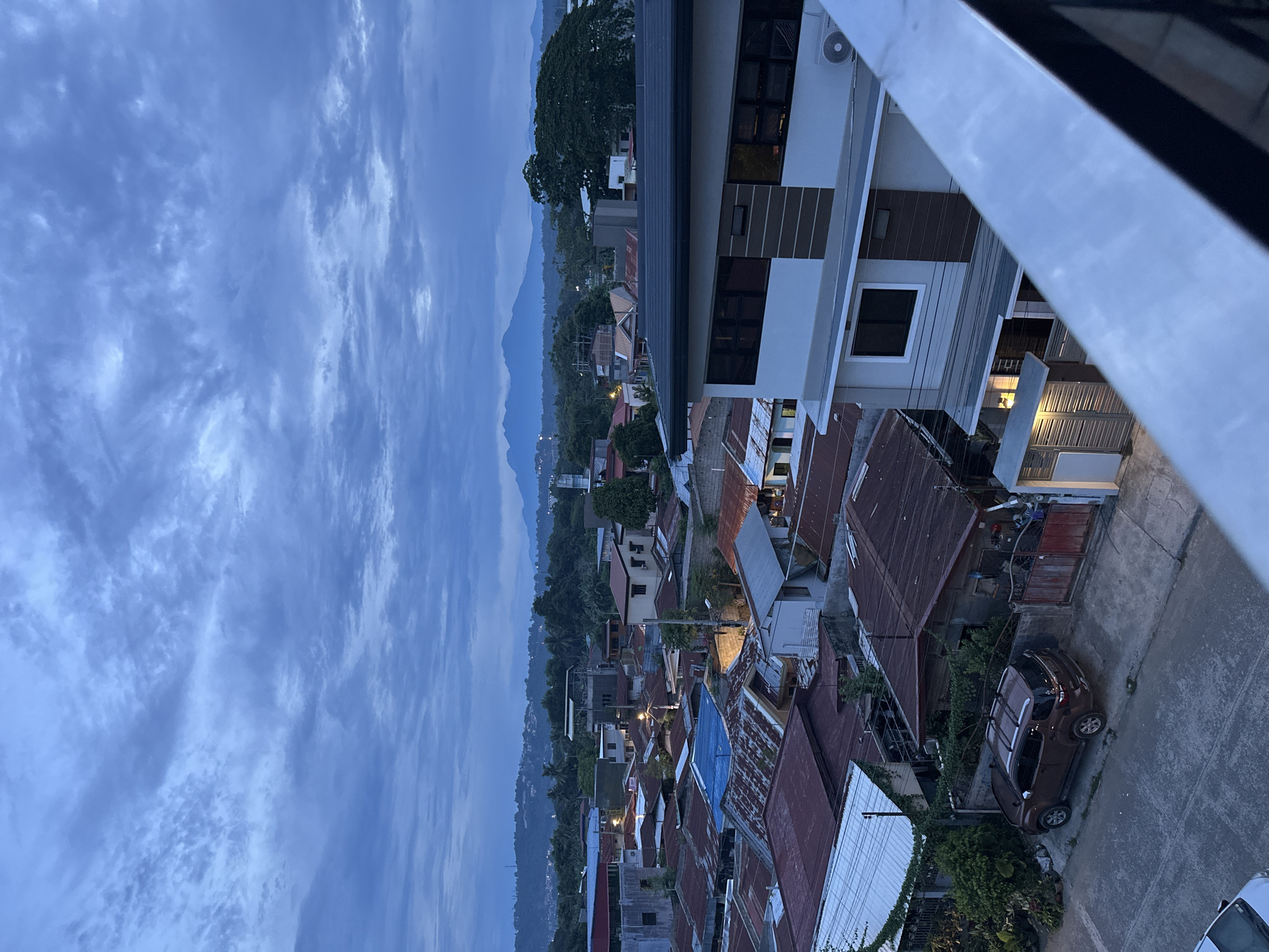 The image shows an elevated view of a section of Davao City, Philippines, at dusk. Mountains are visible in the distance, partially obscured by clouds. The foreground contains buildings, including a modern multi-story structure with light-colored exterior walls. Below it is a dense area of older residential houses with rusted corrugated iron roofs. A brown SUV is parked on a street next to a house with a corrugated iron roof. Streetlights illuminate the narrow streets within the residential area. The sky is overcast with dark blue-gray clouds. A portion of a metal railing or balcony is visible in the upper right corner of the frame.