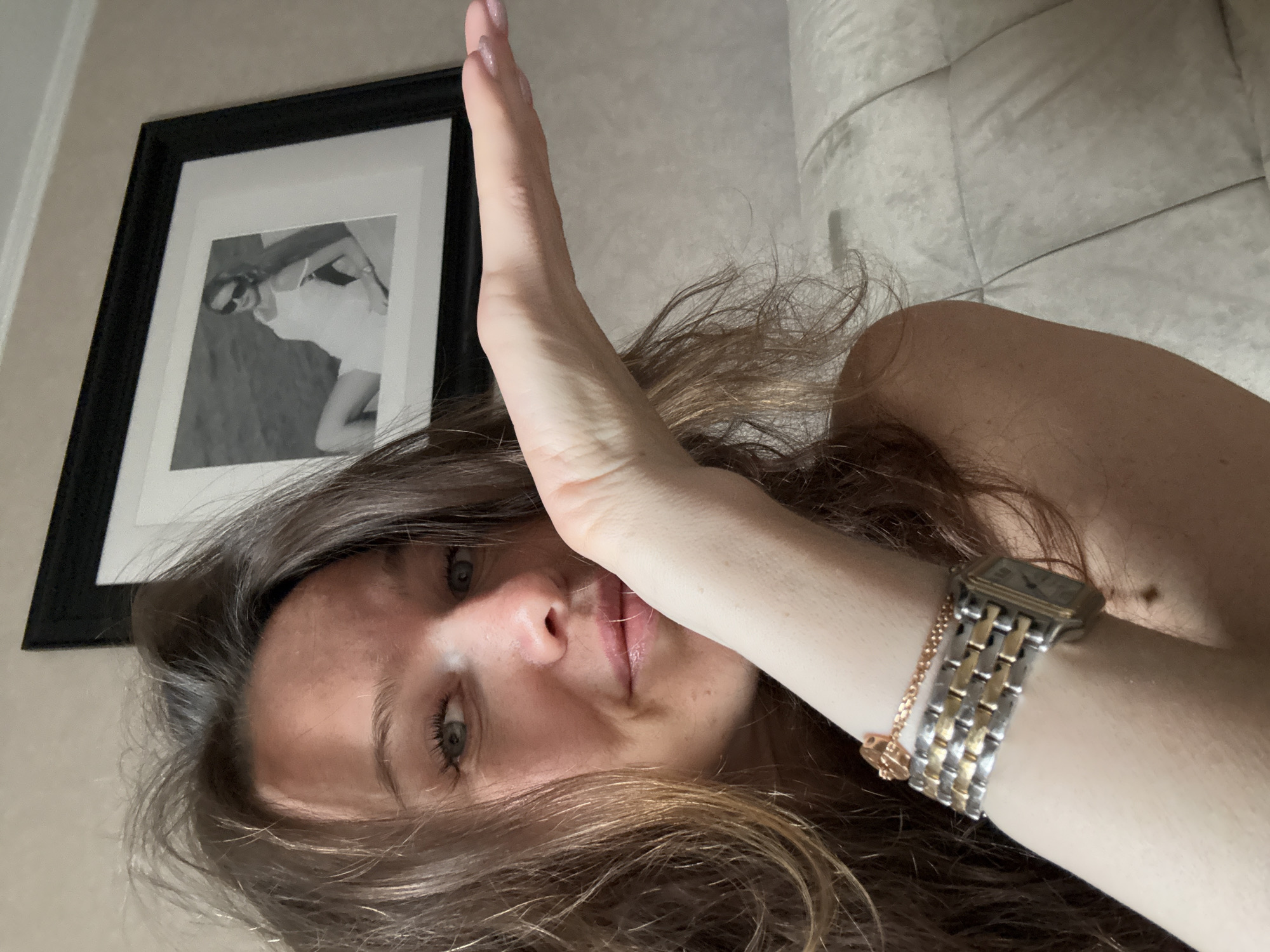 The image presents a close-up, overhead view of a young woman indoors, with her head tilted to the side. She has long, dark brown hair that cascades around her face and shoulders, fair skin, and light-colored eyes (blue/grey). Her right arm is raised, with her hand partially obscuring the upper part of her face. She looks directly towards the camera with a neutral to slightly smiling expression.
On her right wrist, she wears a two-toned metallic wristwatch with a square, dark face, accompanied by a thin, gold-toned chain bracelet with a small, circular charm. Her fingernails are visible with a light, possibly pink, polish.
The background features a framed black-and-white photograph hanging on a light-colored wall. The photo within the frame depicts a woman in a light-colored outfit and sunglasses, posing. To the right and behind the woman's shoulder, a section of a light-colored, possibly off-white or light grey, upholstered surface with a quilted or tufted pattern is visible, suggesting a headboard or a large piece of furniture. The lighting is soft and ambient, characteristic of an indoor setting. The image conveys a casual, personal moment, possibly a selfie. No visible text is present.