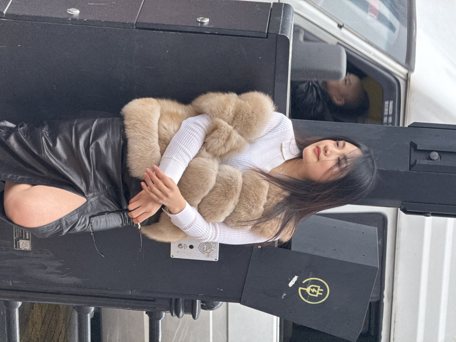 An image, likely taken in Yuzhong District, China, features a young Asian woman posing outdoors. She has long, dark hair that falls over her shoulders and is wearing a voluminous, light beige fur vest over a white, ribbed long-sleeved top with a collar. A black leather skirt or shorts are visible below her waist. Her hands are gently clasped together in front of her, and she has a calm, slightly introspective expression, looking towards the left side of the frame.
The woman is positioned next to and partially leaning on large, dark, rectangular structures, which appear to be street furniture, utility boxes, or possibly parts of a vehicle's exterior. In the background, a white vehicle, resembling a bus or truck, is visible. A reflection of another person, possibly a man, can be seen in the vehicle's side mirror or window.
Notable details include a distinct yellow circular logo on one of the black structures, featuring a stylized electrical plug or charging symbol. Another section of the black structure shows a small, circular metallic grille, possibly a speaker or intercom component. Partially obscured text or a QR code is also visible on one of the black surfaces near the woman's leg. A dark metal railing is present at the bottom edge of the image. The scene is illuminated by soft, diffused daylight, suggesting an overcast day or shaded location.
