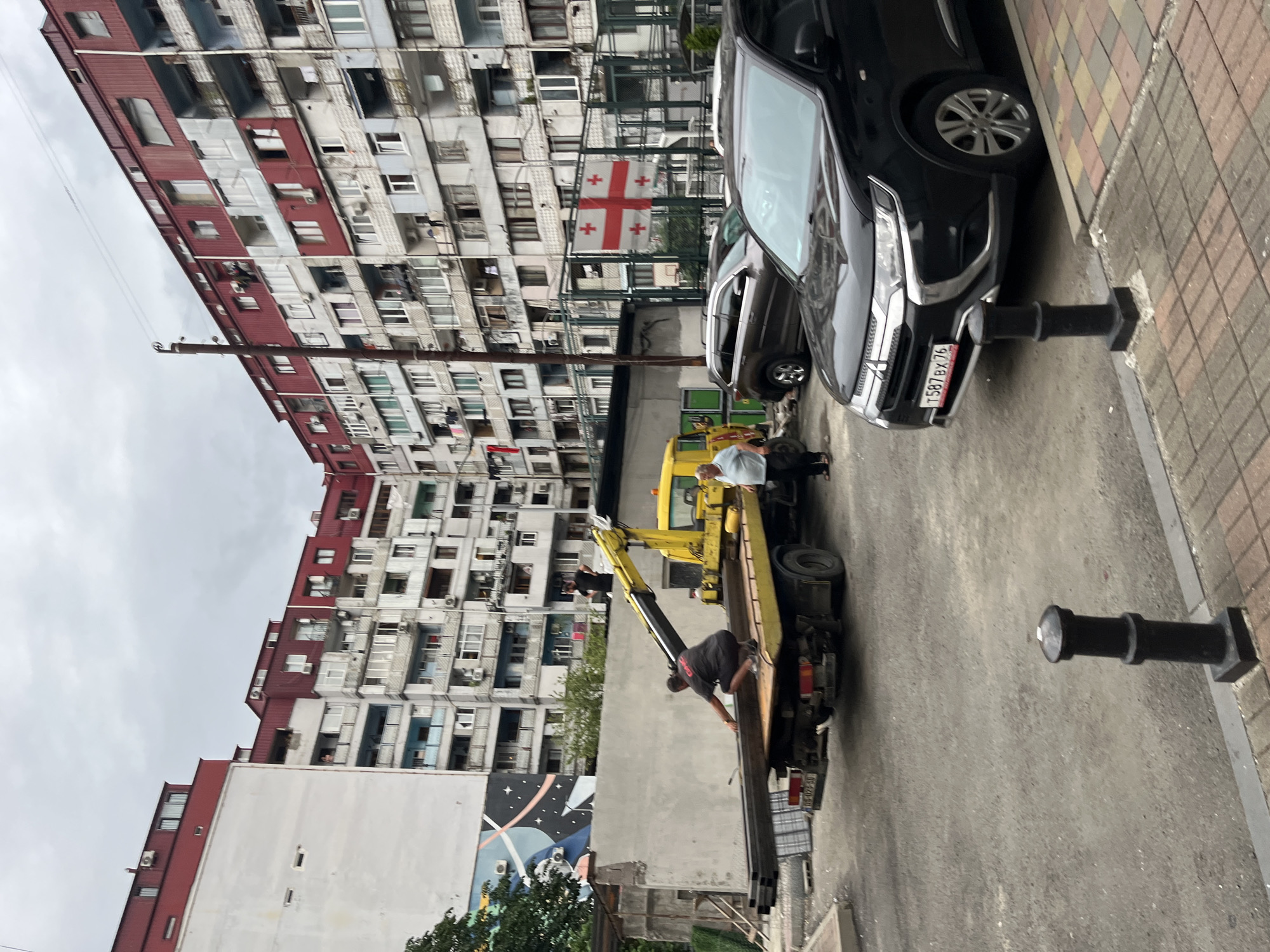 In Batumi, Georgia, on a cloudy day, a yellow tow truck with a crane is parked on a gravel surface, facing right. A man in a grey t-shirt is crouching on the truck bed, arranging metal pipes. Another man in a blue shirt stands on the ground, observing. In the background, a multi-story apartment building with red and white facades dominates the skyline. A Georgian flag is visible on a fence. Several cars are parked in front of the building, including a black SUV in the foreground.
