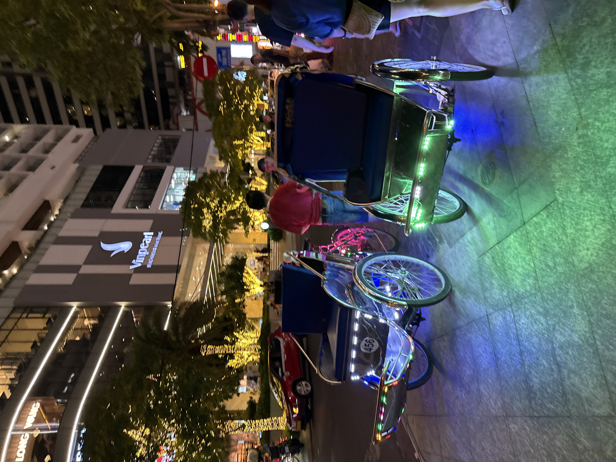 The image captures a nighttime street scene in Nha Trang, Vietnam, likely near a commercial area. Two illuminated cyclos, a common mode of transport in Vietnam, are parked on a tiled sidewalk. The cyclos are decorated with colorful LED lights, predominantly blue and green, casting vibrant reflections on the ground. The one on the left has a visible license plate with the number "456".
In the background, a modern building complex is visible, adorned with numerous string lights that illuminate the trees and facades. A prominent sign displays the "Vinpearl" logo and "BEACHFRONT NHA TRANG", indicating a resort or hotel. Streetlights and building lights contribute to the overall illuminated atmosphere of the urban environment.
Several people are present in the scene. A person wearing a red shirt and blue jeans stands near the left cyclo, possibly a driver or a tourist. Other individuals are seen walking on the sidewalk, some blurred due to motion. A red car is parked on the street behind the cyclos, and other vehicles are dimly visible in the distance. The setting suggests a lively tourist area with ongoing commercial and pedestrian activity.