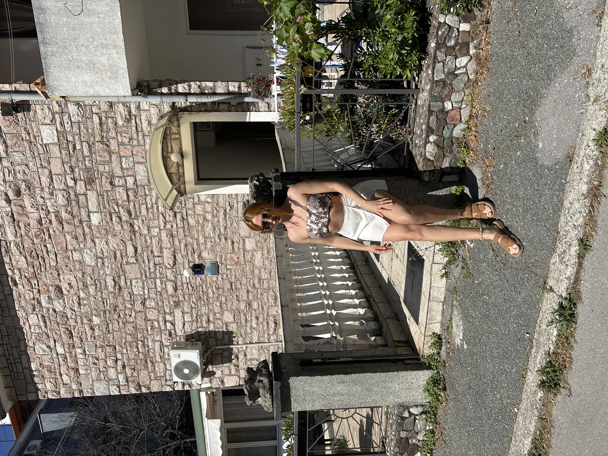 The image depicts an outdoor scene in Budva, Montenegro, on a sunny day. A young adult female, positioned slightly off-center to the right, is leaning against a textured grey stone pillar. She has reddish-brown hair and wears sunglasses, a strapless, patterned crop top, a white mini-skirt, and brown flat sandals with a beaded anklet on her right ankle. Her left arm rests on the pillar, which is topped with an irregular grey stone formation.
Behind the woman and extending across the frame is a building with a facade constructed from rectangular cut stone blocks in various shades of reddish-brown and grey. On the left side of the building, an exterior air conditioning unit labeled "FOX" is mounted on the stone wall. To its right, a blue street number plaque displaying "1" is visible, with a smaller sign below it reading "APARTMAN." A curved, light-colored awning shelters the main entrance door of the building, located directly behind the woman. A white baluster railing runs along a tiled pathway leading to this entrance.
To the immediate right of the woman, a black metal fence delineates a garden area containing various green plants, including a prominent vine with large leaves. Further right, a section of the building features a white stucco finish. A metal downspout runs vertically down the building's facade. In the background on the far left, a bare-branched tree is visible against a darker building or structure. The ground beneath the woman and extending towards the viewer consists of a dark grey aggregate sidewalk, bordered by a concrete curb separating it from an asphalt street. Patches of weeds and grass grow along the edges of the sidewalk and curb. Bright sunlight casts distinct shadows across the scene.