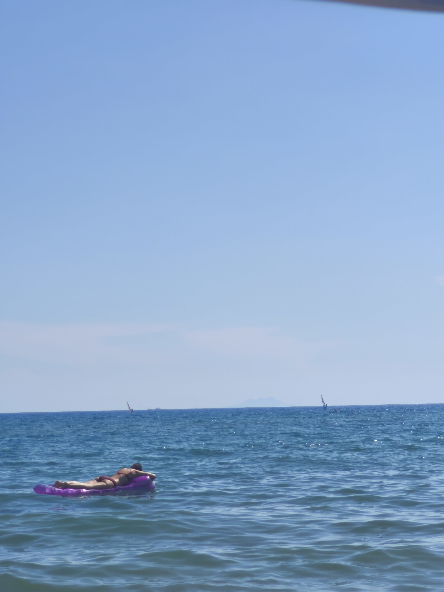 The image depicts a serene seascape in Castiglione della Pescaia, Italy. A person is seen relaxing on a purple inflatable raft, floating on the calm blue water. The person is positioned face down on the raft, appearing to be sunbathing. In the distance, several sailboats are visible on the horizon, indicating a breezy day suitable for water sports. The sky is a clear, bright blue, with a faint haze near the horizon. The overall scene conveys a sense of tranquility and leisure, characteristic of a coastal vacation spot.