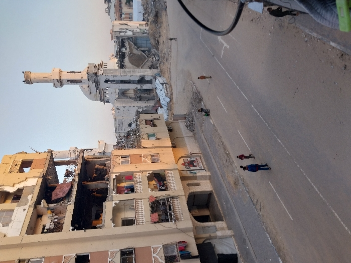 This image presents a scene of extensive urban destruction in Khan Yunis, Palestinian Territory, under a clear sky. On the left, a multi-story residential building displays severe structural damage, with multiple floors featuring collapsed sections, exposed interior rooms, and broken balconies from which clothing is visible. To the right, the main structure of a large mosque is heavily damaged, with its dome significantly compromised and large amounts of rubble accumulated around its base, though its minaret remains largely intact.
A paved road with white lane markings extends through the foreground and mid-ground. Three children are visible on this road: one appears to be running, and two others are walking or standing nearby. Further down the street, on the right, an adult figure stands amidst debris near the mosque, and another individual is partially visible in the extreme foreground on the right, obscured by an overhead cable and a railing. The overall environment is characterized by widespread architectural wreckage and debris.