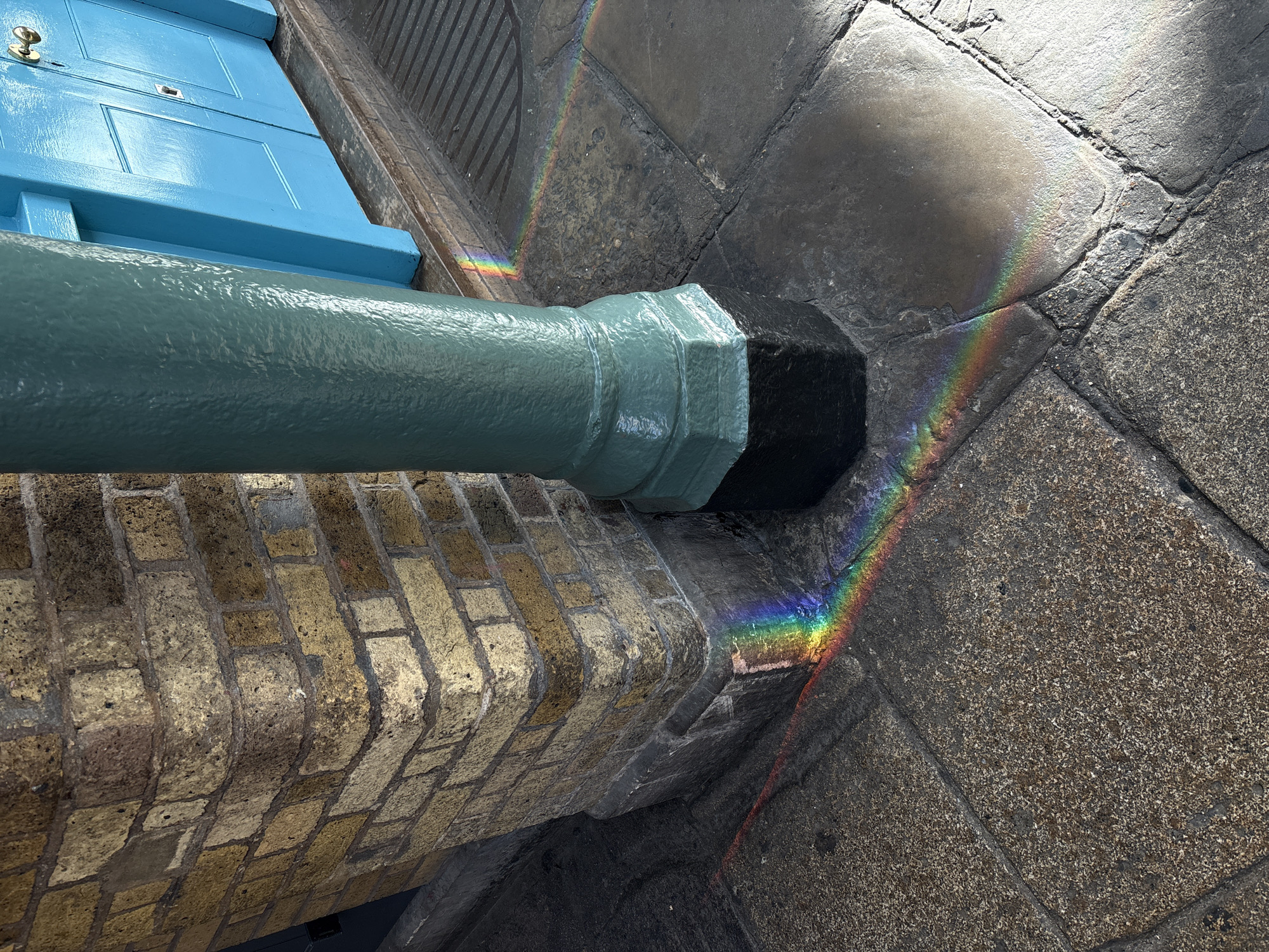 The image captures a close-up view of architectural details in what appears to be London, United Kingdom. A light blue painted door is visible in the upper left corner. A cylindrical, teal-colored metal pipe runs diagonally across the frame, intersecting with a brick arch. The pipe has a black-painted joint. A section of gray stone paving is visible. Refracted light creates a rainbow effect on the stone paving near the pipe and also on the wall next to the door.