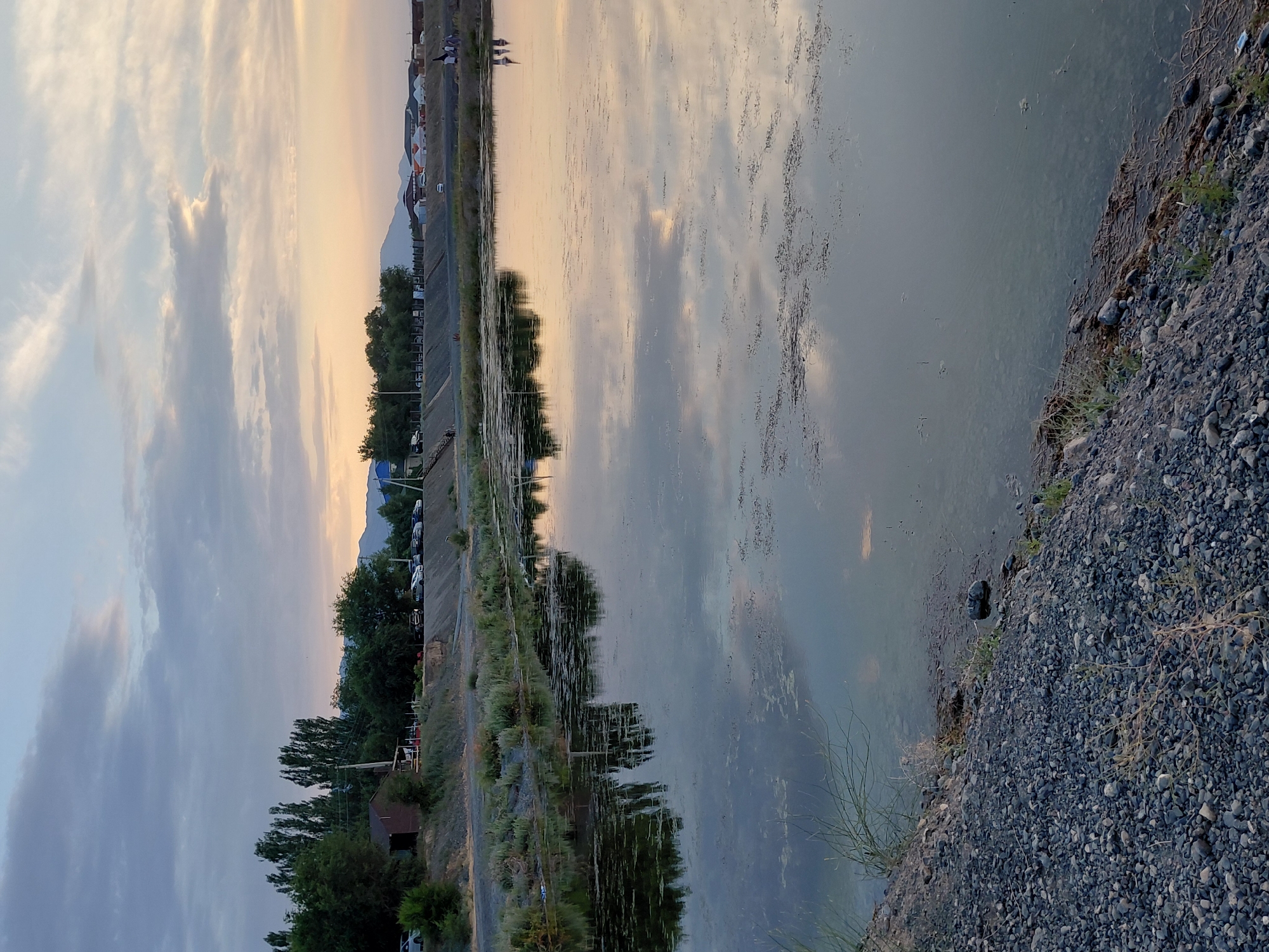 The image depicts a scenic view of a body of water, likely a lake or reservoir, at dusk in Ақши, Kazakhstan. The sky is a soft gradient of pastel colors, with wispy clouds reflecting on the calm water's surface, suggesting an early evening or late afternoon setting. The water itself appears slightly murky with some organic debris floating on it.
On the left side of the image, a well-maintained embankment runs along the water's edge, separating it from a road and a small settlement. The embankment is lined with concrete structures and vegetation, including trees and bushes. Further inland, a row of cars is parked along the road, and in the distance, some buildings and yurts can be seen, hinting at a residential or recreational area. A few people are visible near the water's edge, possibly enjoying the twilight.
The right side of the image shows a rocky shoreline with sparse vegetation, composed of small grey pebbles and dry grass. The overall atmosphere is tranquil and serene, with the fading sunlight casting a warm glow on the landscape. The natural beauty of the scene is complemented by the human elements that suggest a peaceful community life by the water. No visible text is present in the image.