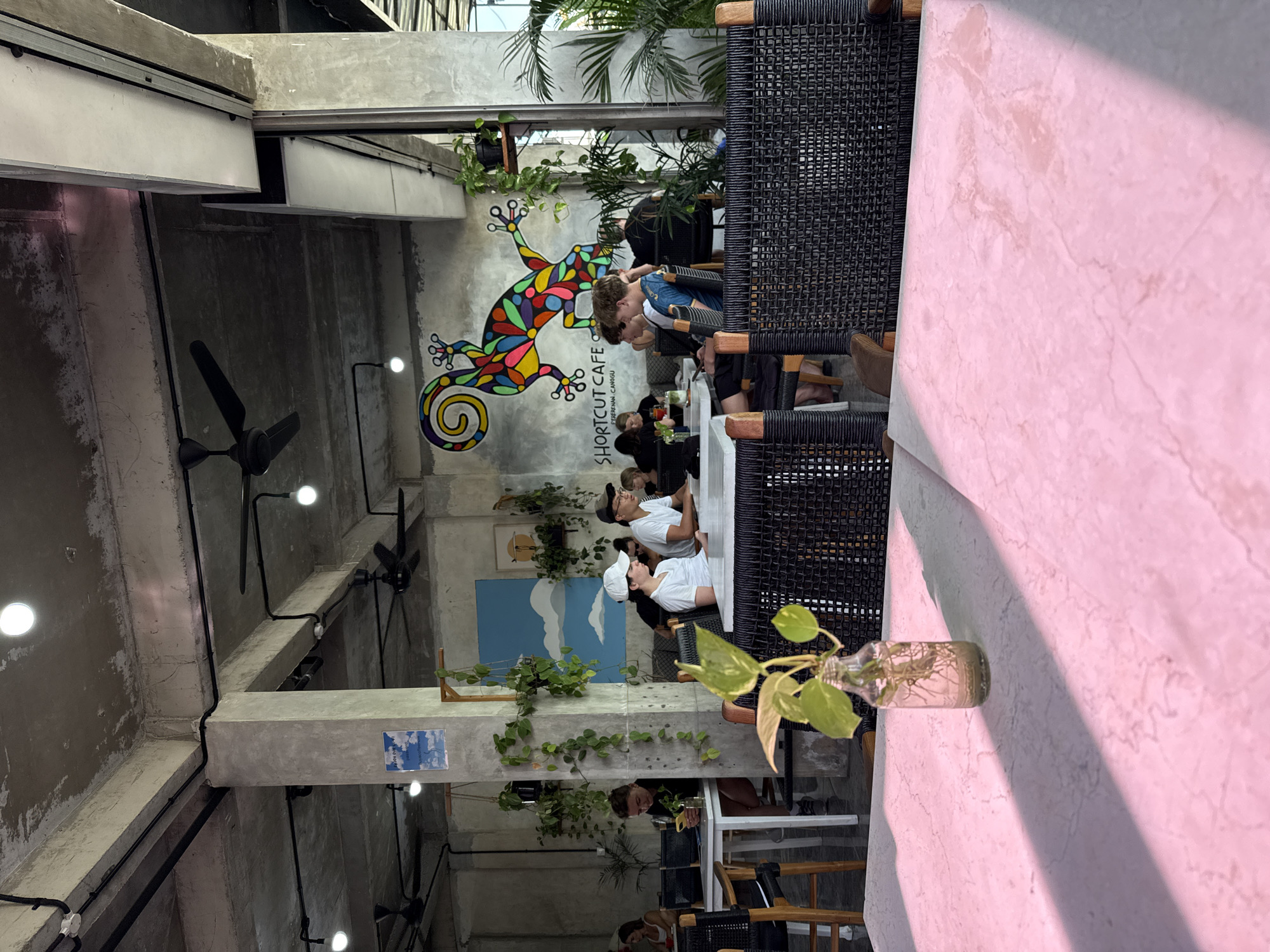 The image shows the interior of the Shortcut Cafe in Canggu, Indonesia. The interior features a mural of a brightly colored lizard, exposed concrete beams and walls, hanging plants, and overhead fans. Several people are seated at tables, some looking at their phones, others conversing. The tables have woven black chairs. A small glass bottle containing a plant sits on the edge of a pink-toned surface in the foreground.