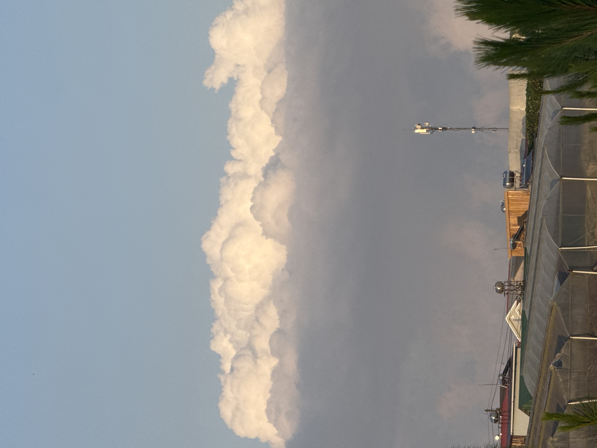 The image captures a scene in Lâm Viên - Đà Lạt, Vietnam, featuring a vast expanse of sky dominated by a thick band of luminous clouds illuminated by sunlight. Below the clouds, a lower layer of grayish, diffused clouds partially obscures the landscape. The foreground is occupied by several structures including greenhouse-like buildings with translucent coverings, some residential rooftops with water tanks, and a communication tower equipped with antennas. Green foliage, likely from pine trees, is visible in the lower right corner. The overall atmosphere suggests an outdoor environment, possibly in the early morning or late afternoon, given the quality of light.