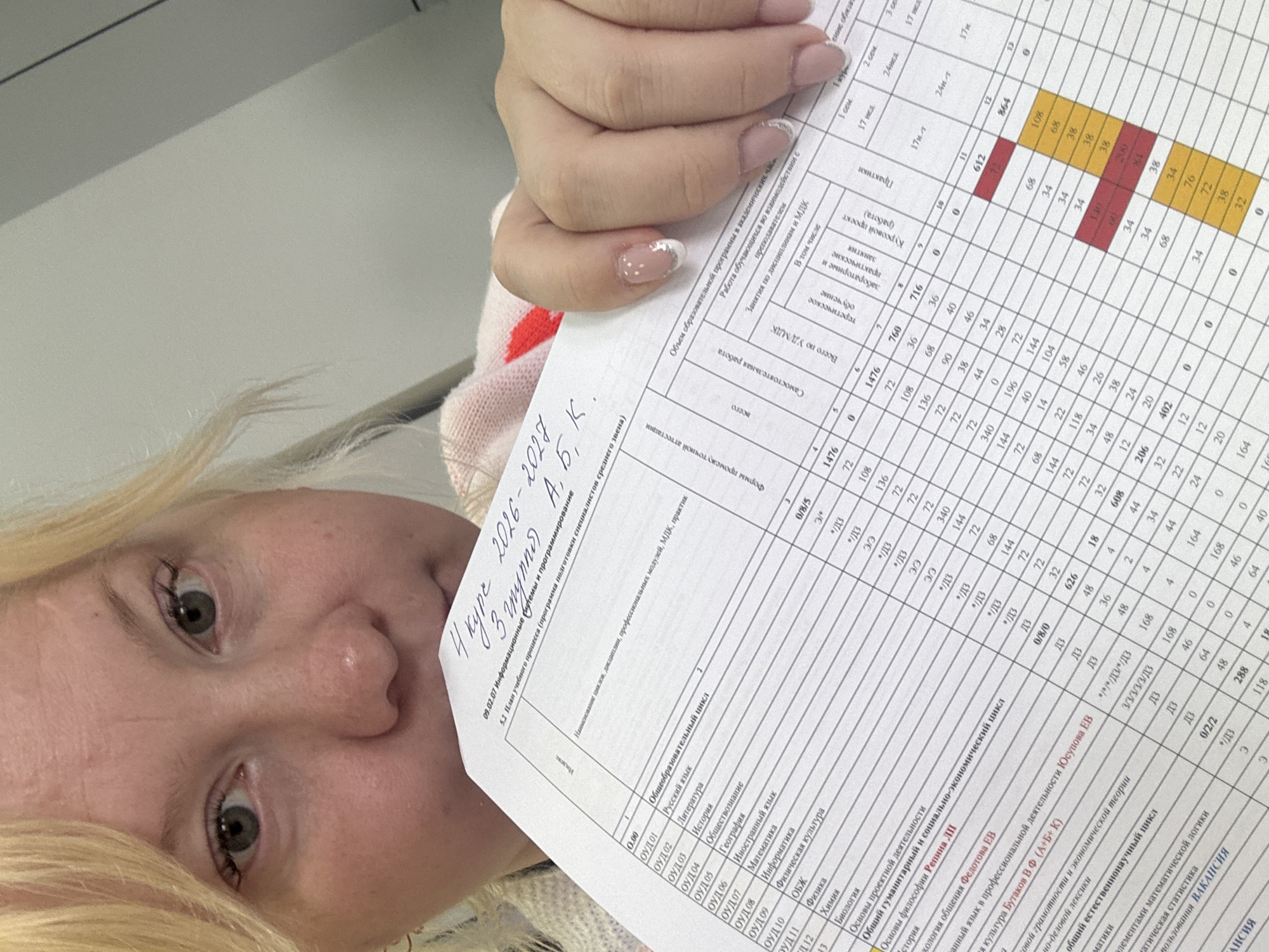 Here is a description of the image:
**People:** A person with fair skin and blonde hair is holding a piece of paper. Their eyes are visible and appear to be looking up and to the left. The person has manicured nails.
**Subjects:** The main subject of the image is a document filled with a table of data. The document seems to be related to an academic curriculum.
**Objects:** The objects in the image include a piece of paper, which seems to be a printed academic document or a schedule, a hand, and the person's face and upper body.
**Actions:** The person is holding the paper up, likely for viewing or reading.
**Interactions:** The person interacts with the document by holding it up for view. The document contains information related to course curriculum.
**Environment:** It's difficult to determine a specific location. The background is blurred and does not offer significant environmental details. Based on the text in the document, the location is likely in Russia. It is impossible to determine if the location is the city of Chaikovsky, Russia from the given image.