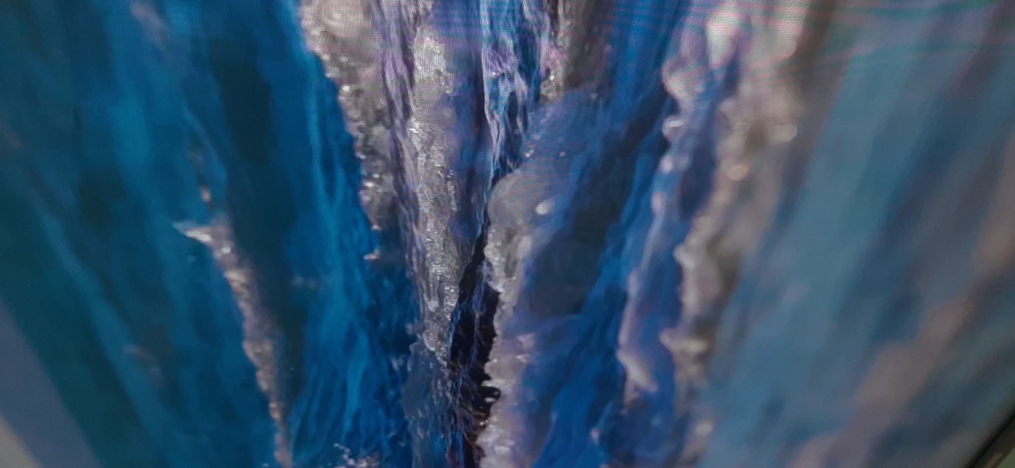 The image is a close-up, slightly blurred photograph of ocean waves crashing. The water is a deep blue with white foam and spray catching the light, creating a shimmering effect. The waves appear to be in motion, with crests and troughs clearly defined. The perspective is low, as if the camera is at sea level or slightly below. There is no indication of any specific location within the city of Balashikha, Russia, as the scene is purely natural and devoid of any urban or identifiable geographical features.