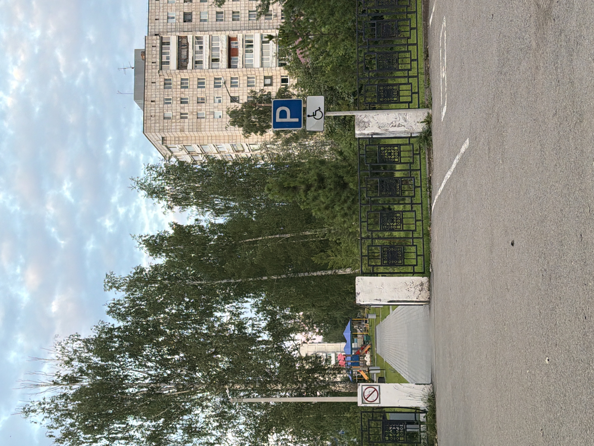 The image captures an outdoor scene in the city of Chaikovsky, Russia. In the foreground, there is a paved surface with white markings indicating a parking spot for individuals with disabilities. A black metal fence with decorative elements runs horizontally across the middle ground. To the left, a white pillar supports a circular sign with a red border and a diagonal line through a cigarette, signifying "No Smoking." Adjacent to this pillar is an open gateway leading to a paved walkway.
Beyond the walkway, a children's playground is visible, featuring brightly colored slides and play structures under blue canopies. Tall, mature trees with lush green foliage frame the playground and walkway. In the background, a multi-story apartment building with a light-colored, textured facade stands prominently against a cloudy sky with patches of blue. A street lamp is positioned near the apartment building. The overall environment suggests a residential area with recreational facilities.
