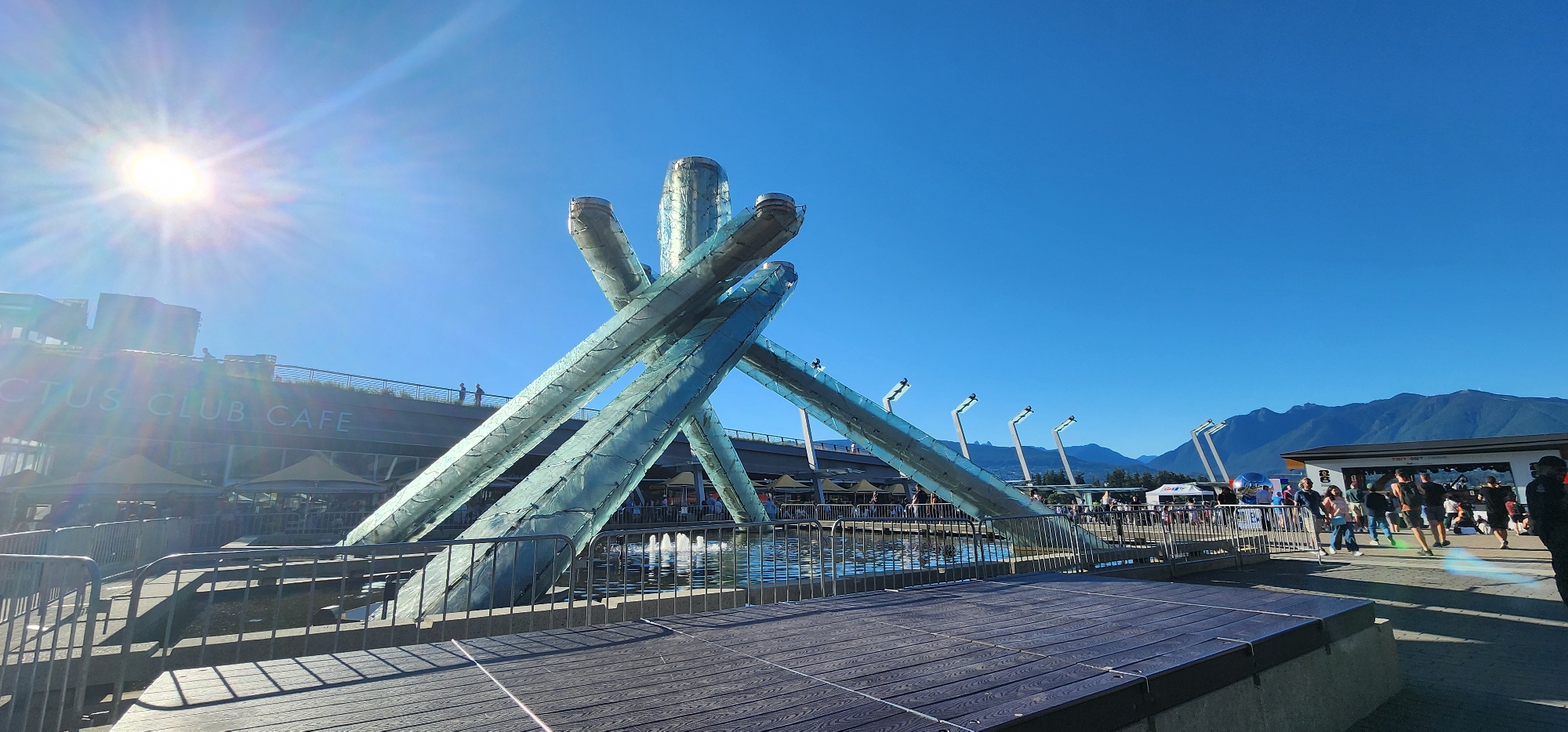 The image shows a wide shot of the Olympic Cauldron at Jack Poole Plaza in Vancouver, British Columbia. The sun is shining brightly, casting lens flares across the sky and the sculpture. In the background, there is a building with "CACTUS CLUB CAFE" written on it, along with several outdoor umbrellas. People are visible walking around the plaza, and mountains can be seen in the distance. The Cauldron itself is a large, abstract sculpture made of glass and metal. In the foreground, there is a wooden deck with metal railings. The sky is a clear blue.