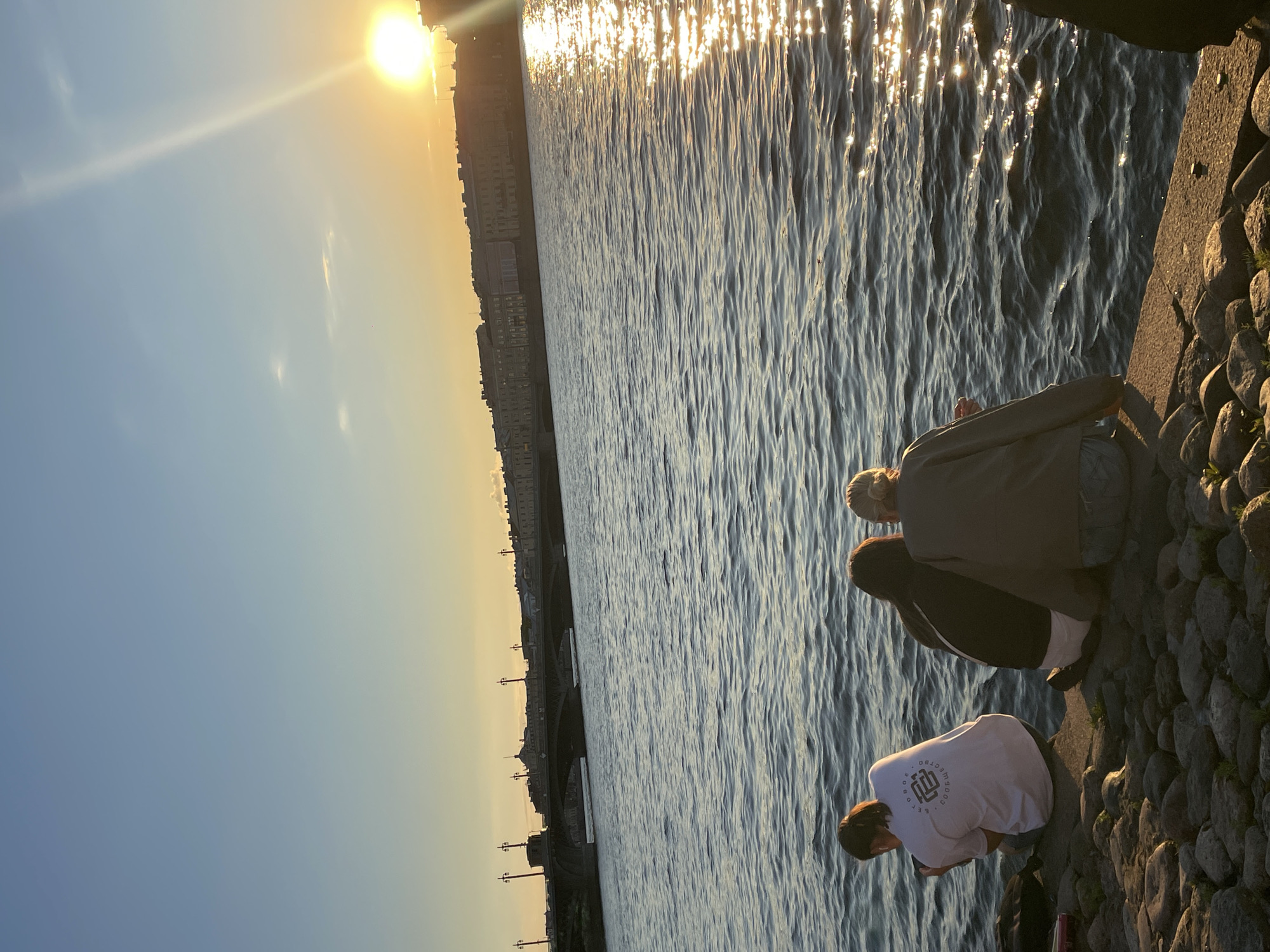The image depicts a waterfront scene in Saint Petersburg, Russia, during sunset. Three individuals are seated on a stone embankment overlooking a body of water. In the background, a bridge with lampposts spans the water, and a cityscape with multi-story buildings is visible on the opposite bank. The sun, low on the horizon, casts a bright glare on the water's surface, creating shimmering reflections. The sky is a gradient of blue and yellow, indicative of the setting sun. The overall atmosphere is calm and serene.