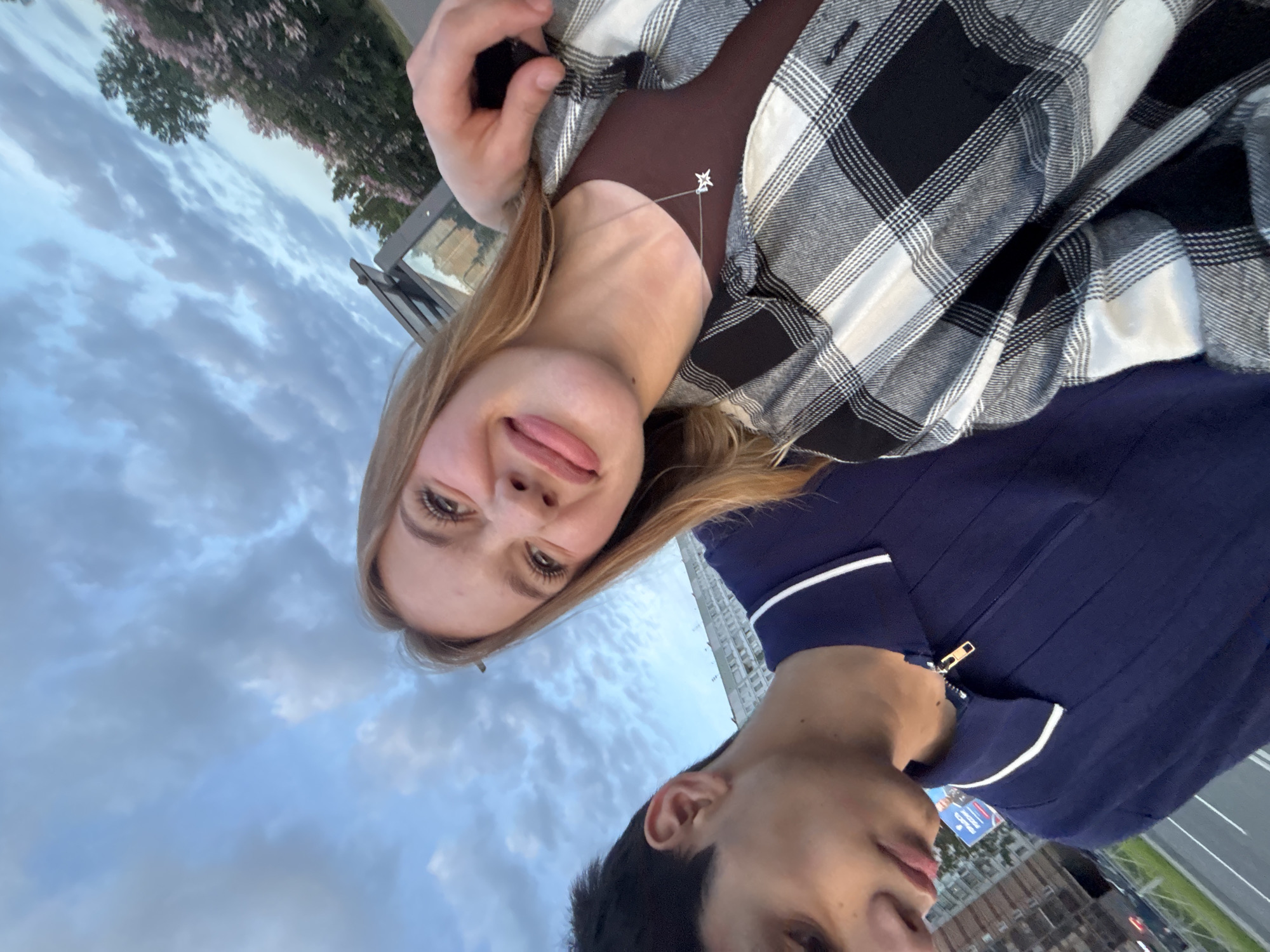 The media file shows a close-up, rotated selfie of two young people, a woman and a man, against a sky with clouds. The woman is in the foreground, her head tilted back, smiling. She has blonde hair and is wearing a brown top under a black and white plaid shirt. A delicate star-shaped necklace is visible on her neck. The man is partially visible in the lower right of the frame, looking away. He is wearing a dark blue shirt with white trim and a zipper.
The background consists of a cloudy sky and a hint of greenery from trees with pink blossoms. In the distance, there appears to be a cityscape with buildings and a road. The lighting suggests it might be late afternoon or early evening. There is no discernible text within the image. The overall mood appears casual and happy.