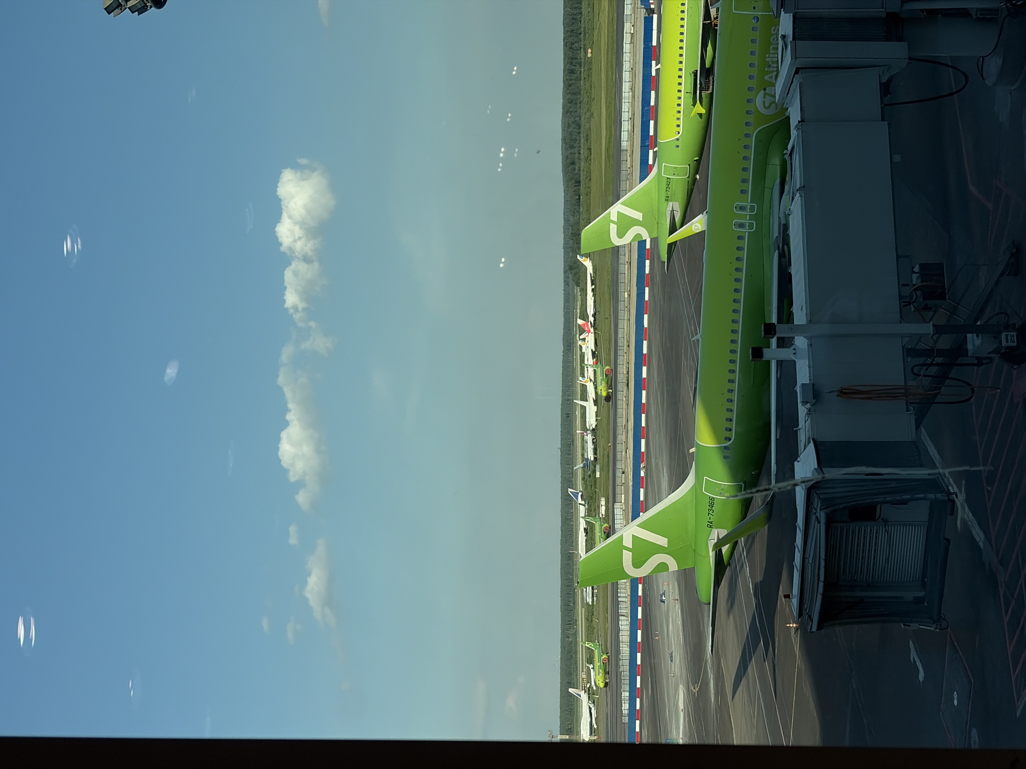 The image displays a daytime view of an airport tarmac and runway area, observed from an interior window of an airport terminal at Domodedovo, Russia. The scene is dominated by multiple aircraft under a clear blue sky with scattered white cumulus clouds.
In the mid-ground, a vibrant green S7 Airlines aircraft is prominently featured. Its fuselage, bearing the registration RA-73423, is partially visible, and the tail fin displays the distinctive white "S7" logo. This aircraft is connected to a gray jet bridge that extends from the unseen terminal structure into the lower-right foreground. Another S7 Airlines aircraft, with registration RA-73466, is positioned behind the first, its tail and part of its fuselage visible. Further in the distance, additional green S7 Airlines aircraft are parked alongside various other airliners, which appear predominantly white.
The tarmac surface is dark asphalt, marked with white lines and red-and-white striped barriers, indicating operational zones. The background consists of a flat, green expanse of land leading to a dense, dark green treeline that forms the horizon. Above the treeline, faint industrial-like structures are visible in the far distance.
The top portion of the sky exhibits several bright, elongated, and circular reflections on the window glass, likely originating from interior terminal lights or structures. No individuals are discernible within the frame. The overall lighting suggests a sunny day.
