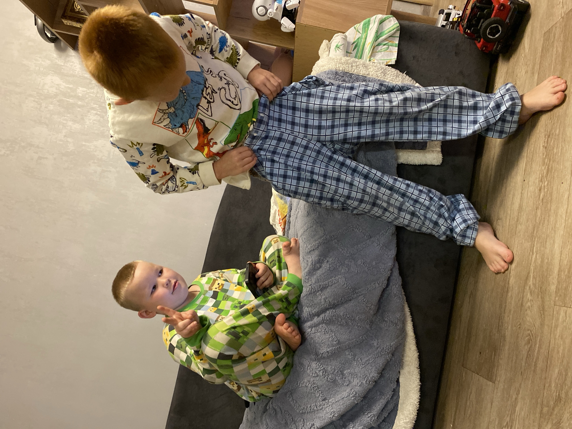 The image depicts an indoor scene featuring two young boys in pajamas. The boy on the left, with blond hair, is seated on a dark gray sofa covered with a blue, textured blanket. He is wearing green and white checkered pajamas and holding a black mobile phone in his right hand, with his left hand raised, displaying two fingers. The boy on the right, with reddish-blond hair, is standing and appears to be adjusting his blue and white checkered pajama pants. He is wearing a long-sleeved pajama top with various cartoon dinosaur illustrations. In the background, a wooden shelf unit is visible, adorned with a small white robot toy and other items. A red toy Jeep is positioned on the floor to the right of the sofa. The floor is made of light-colored wood-grain laminate. The overall environment suggests a residential setting, likely a living room or bedroom. There is no identifiable indication of the specific city of Noviy, Russia within the image itself.