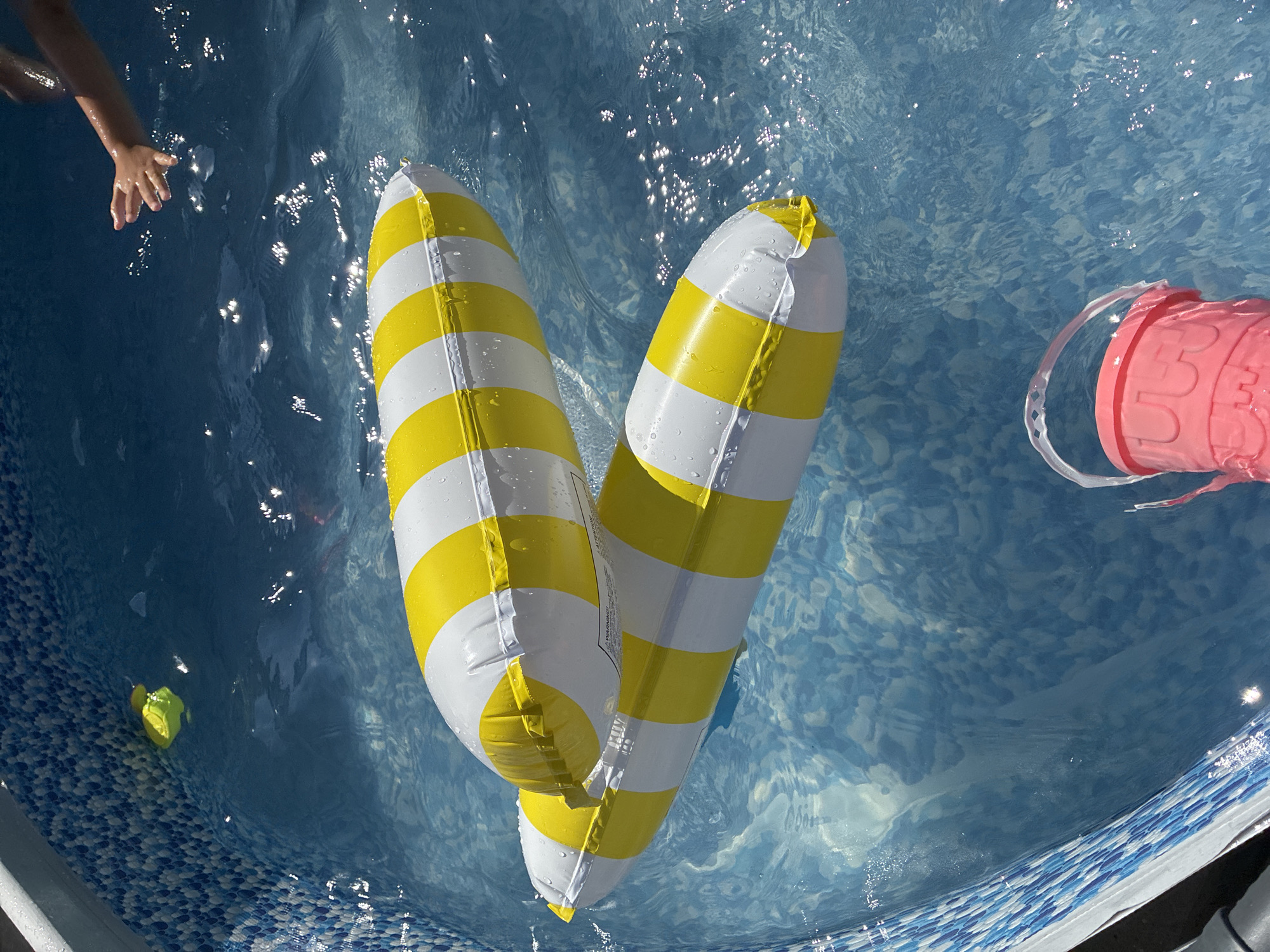 An overhead, close-up view captures two yellow and white striped inflatable pool floats resting on the surface of a blue pool. Sunlight glints off the water's surface, creating shimmering highlights. In the upper right corner of the image, a child's arm and hand reach out towards the floats. In the lower right, a bright pink toy bucket is partially submerged in the water. The pool's interior walls are lined with a blue and white tiled pattern. This scene is taking place in a residential swimming pool in the city of Surami, Georgia.