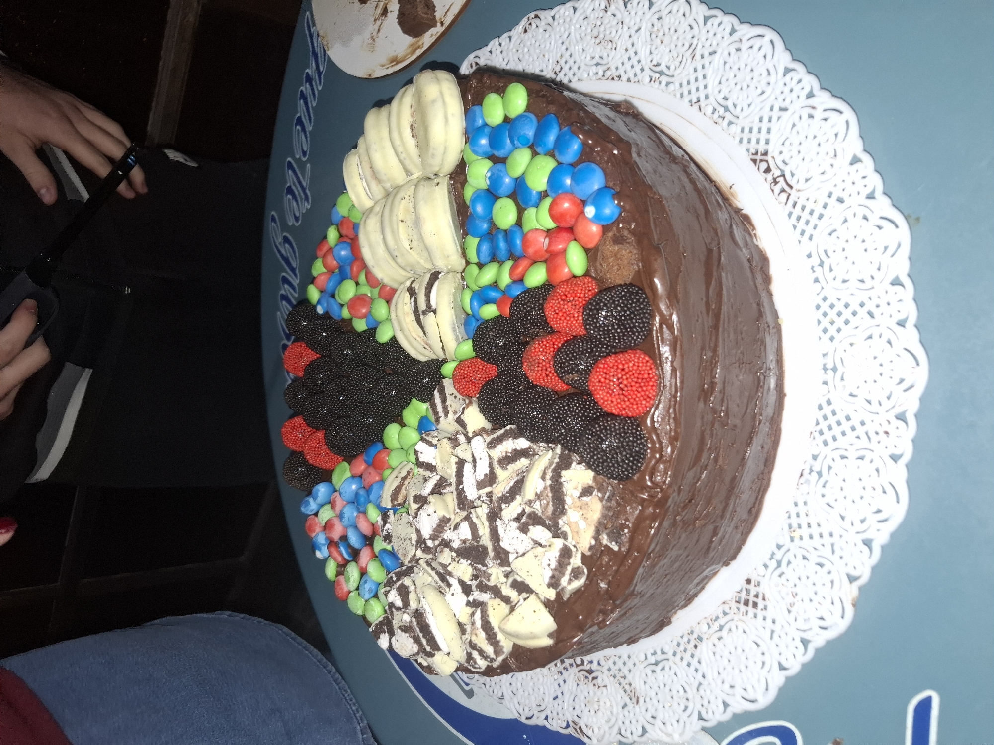 A close-up, overhead shot depicts a decorated chocolate cake on a white lace doily placed on a blue table. The cake is roughly divided into sections. One section is decorated with white macarons, another with multi-colored candies (red, blue, green). A third has black and red gummy candies, and a fourth features crushed cookies and cream, likely Oreos, on a chocolate base. The remainder of the cake appears to be covered in chocolate frosting. Part of a person's arm and blue jeans are visible on the lower left side of the frame. A person's hand holding an object (possibly a lighter or pen) is seen on the upper left. The table has blue and white text. Due to the style of text this is likely located in the city of Buenos Aires, Argentina.