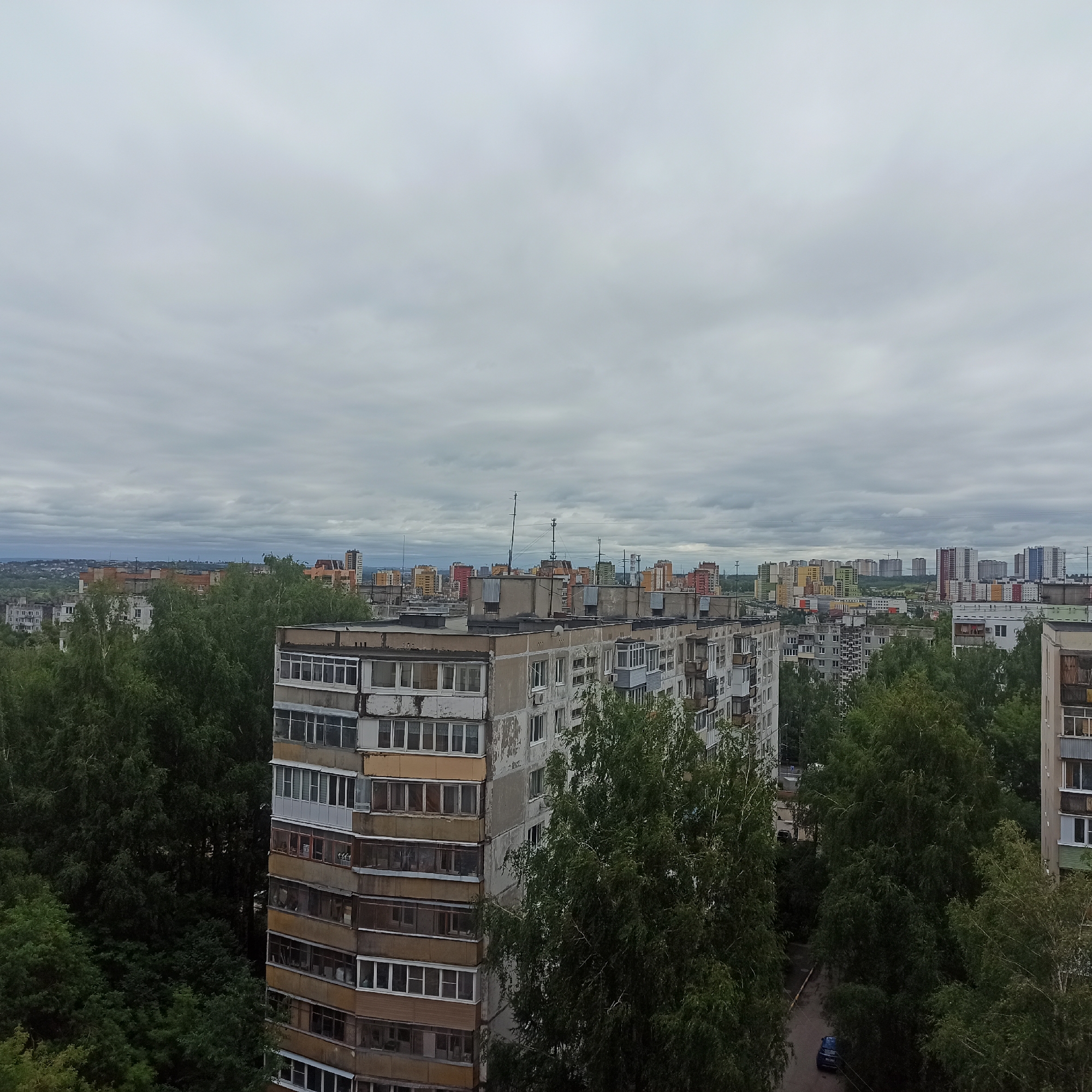 The image depicts a panoramic view of a residential area in Nizhny Novgorod, Russia, under an overcast sky. In the foreground, a multi-story apartment building with a facade of concrete and balconies dominates the scene, featuring windows of various sizes and colors. Dense green trees surround the building, with some foliage extending in front of the lower floors.
Beyond this building, a cityscape unfolds with numerous other apartment buildings, some appearing older and grayer, while others are newer, featuring vibrant colors such as yellow, green, red, and blue. These buildings vary in height and architectural style, creating a dense urban landscape. Antennae and satellite dishes are visible on the rooftops of some structures.
In the distance, the horizon is marked by a lighter haze, suggesting a broader expanse of the city. A few streetlights and utility poles can be discerned among the buildings. A dark blue car is parked on a wet asphalt road visible between the trees and apartment blocks in the middle ground. The overall impression is that of a typical, somewhat weathered, urban environment in a Russian city on a cloudy day.